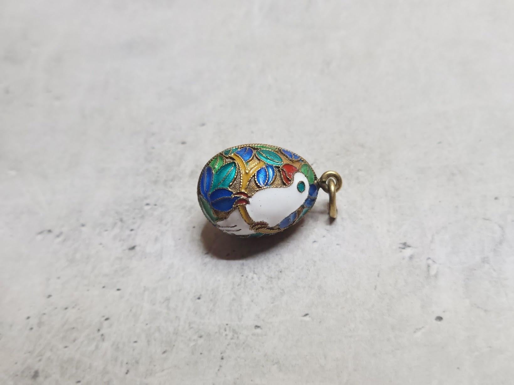 Questo splendido ciondolo a forma di uovo russo in stile Art Déco presenta un vibrante smalto cloisonné che raffigura un pappagallo appollaiato su un ramo, circondato da foglie esotiche. Il disegno dettagliato appare su entrambi i lati dell'uovo,