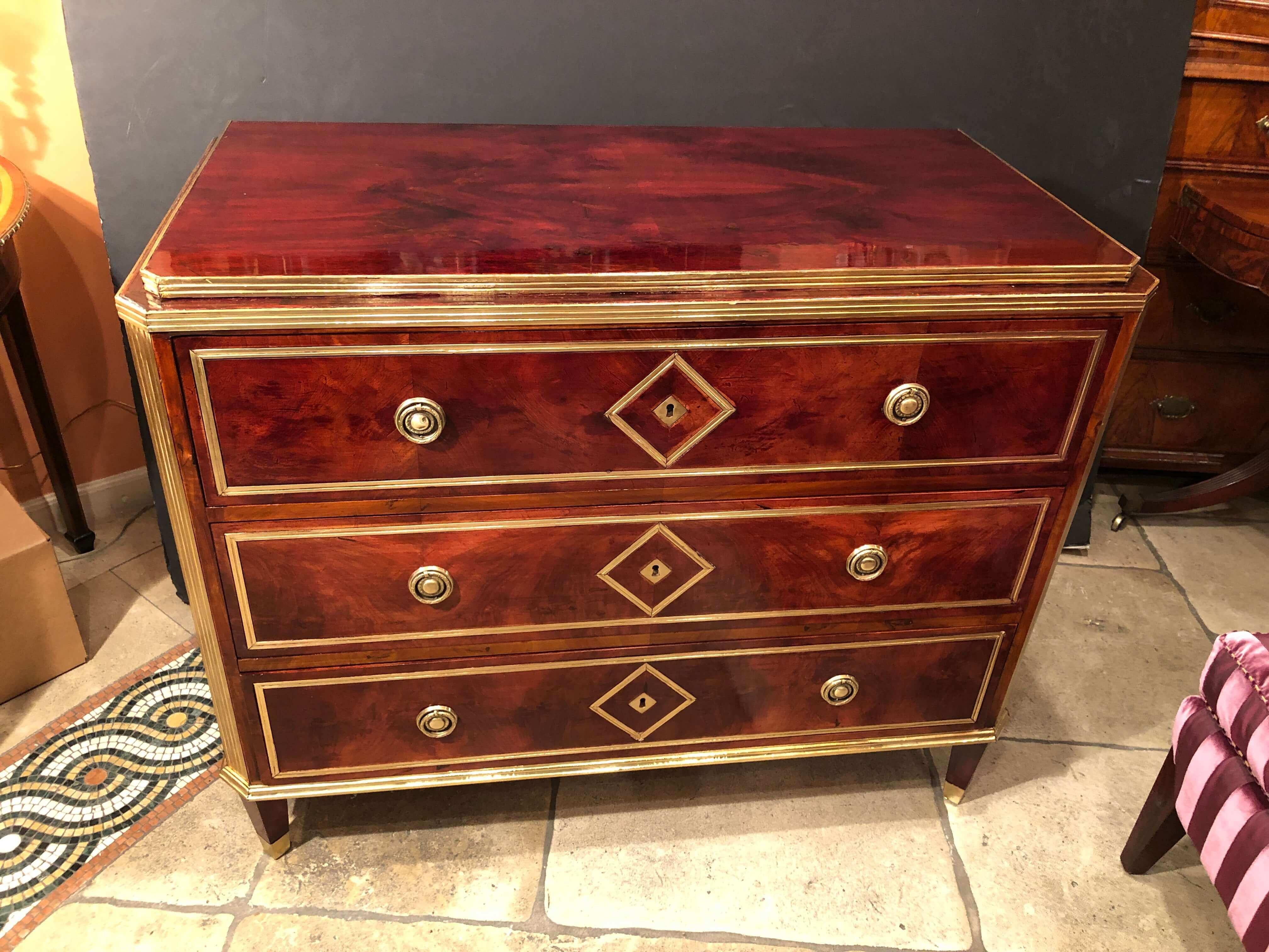 A large Russian neoclassic mahogany three-drawer commode with brass trim and mounts, with diamond motif, canted corners and raised on square tapered legs.