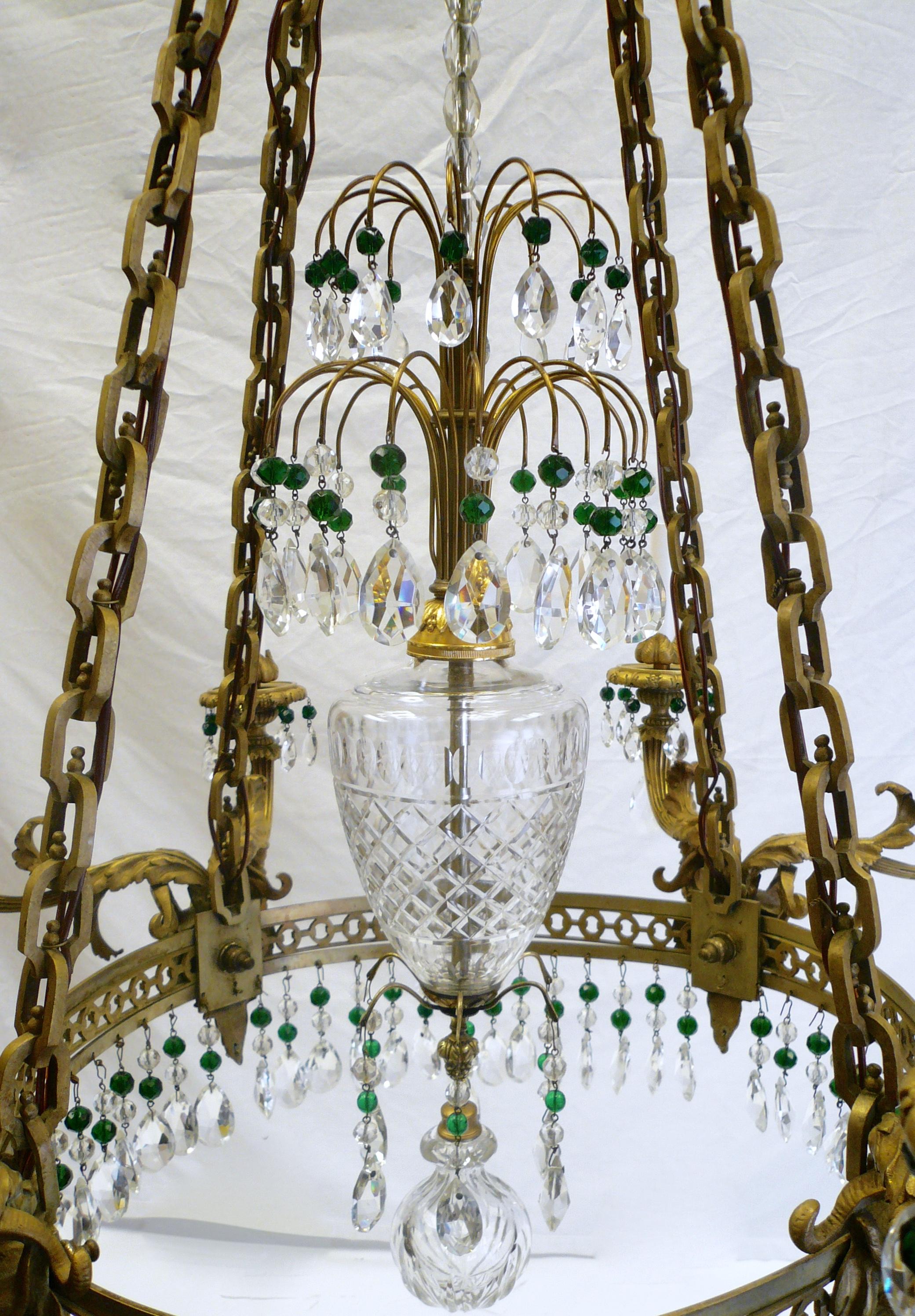 This eight light Alexander I style chandelier features pear shaped faceted crystals, and emerald green crystal accents. The gilt bronze frame features neoclassical motifs including acanthus leaves and rams heads.