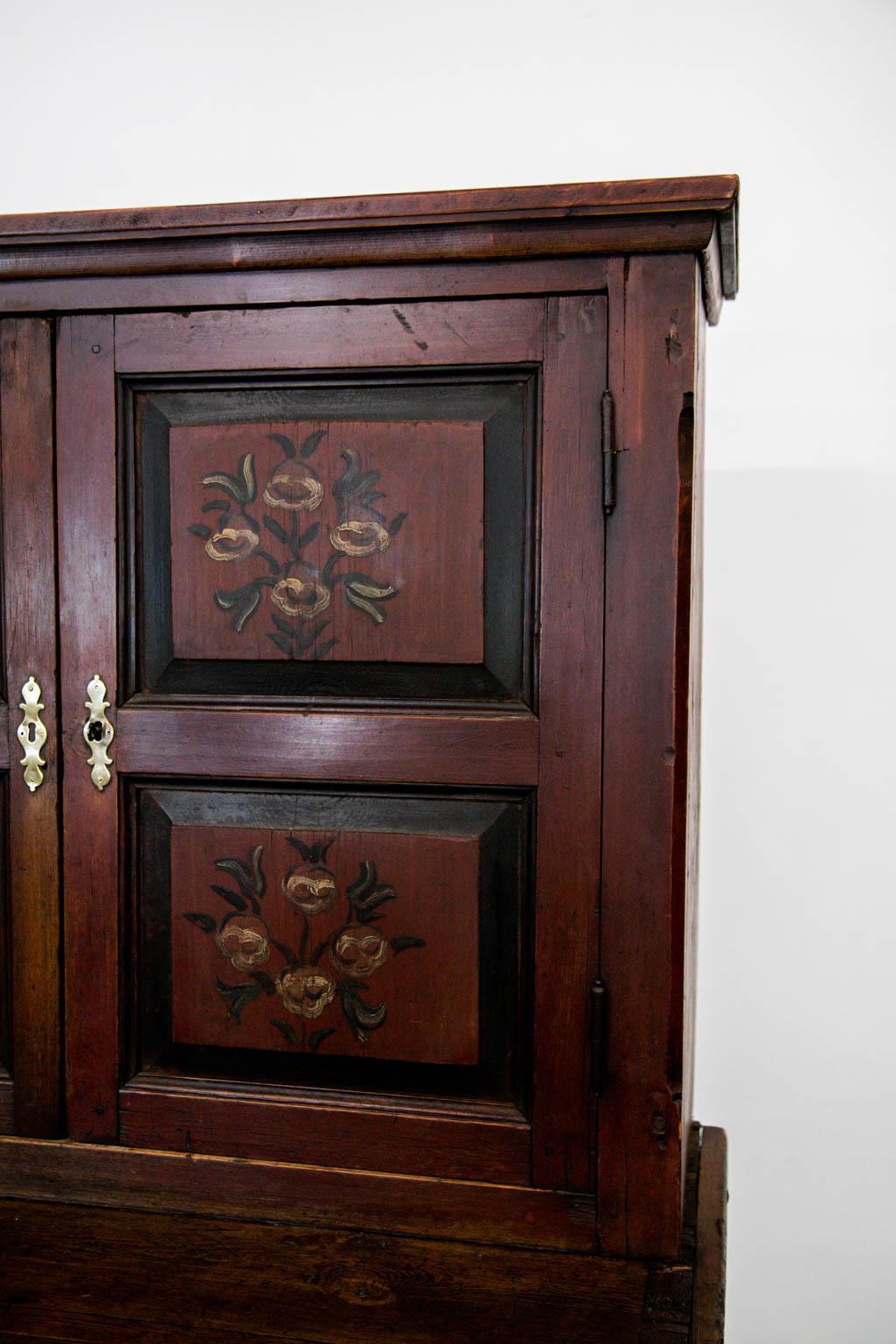 The top doors of this cupboard have four raised panels framed with shaped moldings. The top and bottom doors have a working lock and key. The upper doors have floral painting on the raised panels which have wear commensurate with age and use. There