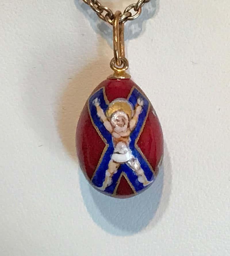 A beautiful porcelain miniature egg pendant, featuring the Cross of Saint Andrew the First Called on a red background. The egg is painted by hand, the reverse with white tipped gilded initials. Attached to a large gilded suspension ring to