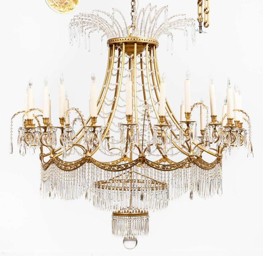 Large twenty-four light basket chandelier in the Russian Empire taste hung with faceted crystal pendants, buttons and beads, having a gold finished frame, curving upper corona, central swaged corona with classical masks issuing arms, with two lower