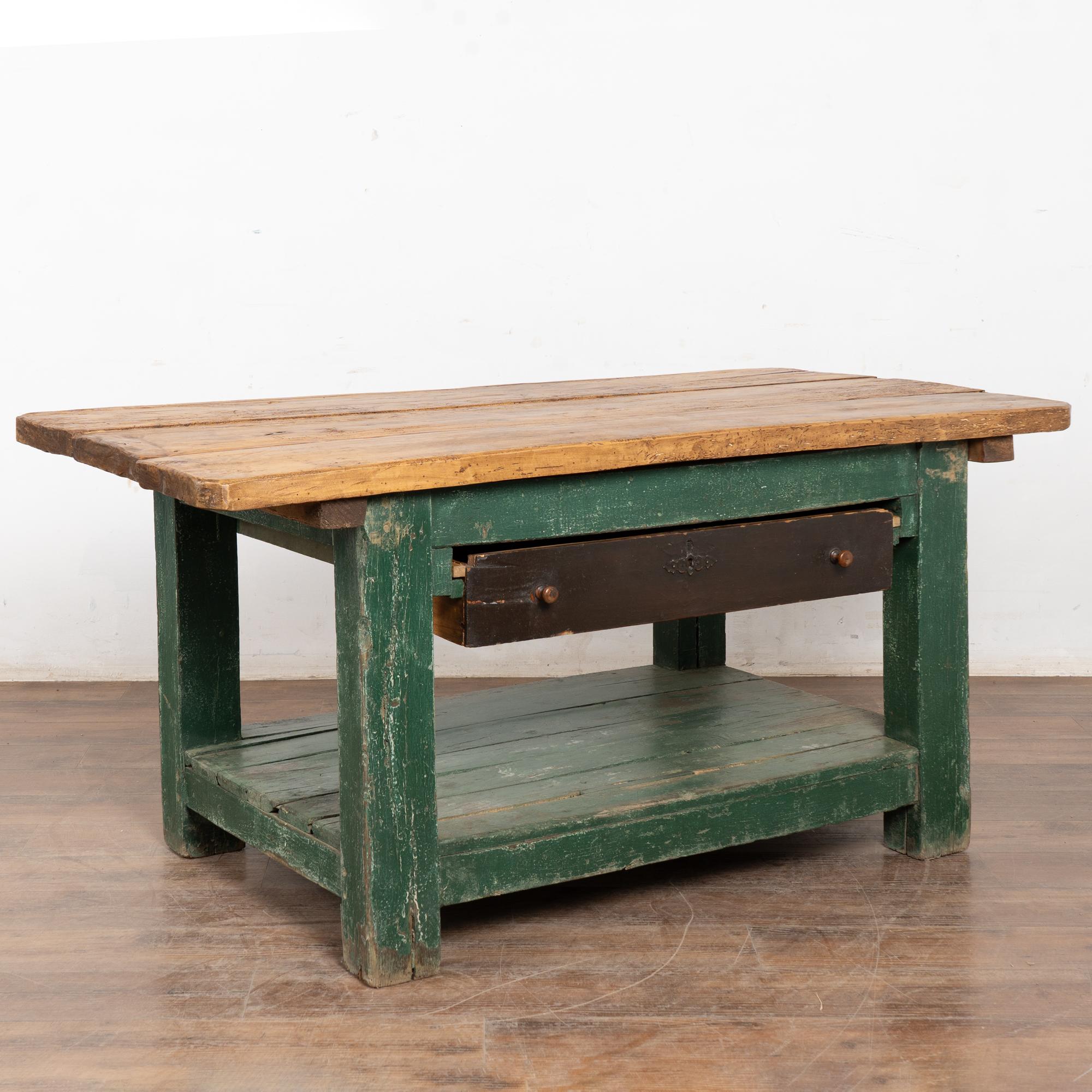 Mesa de trabajo o mostrador de taller rústico con un cajón y estante inferior. El acabado pintado de verde está desgastado en toda la base, mientras que el cajón está pintado de un marrón que contrasta.
Esta mesa de 1,80 m muy usada refleja