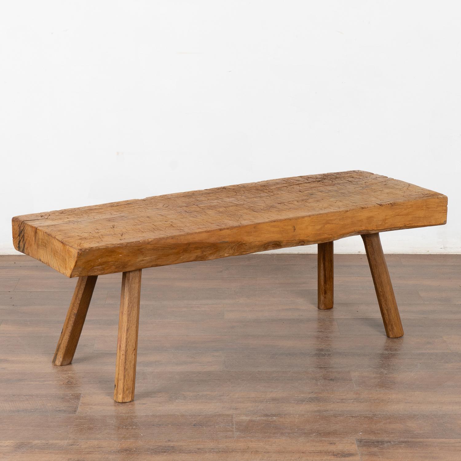 The appeal of this rustic coffee table comes from the thick hard wood top that originally served as a work table, resting on strong peg splay legs. 
Note the heavy gouges, nicks, old cracks and stains that all combine to build the vintage character