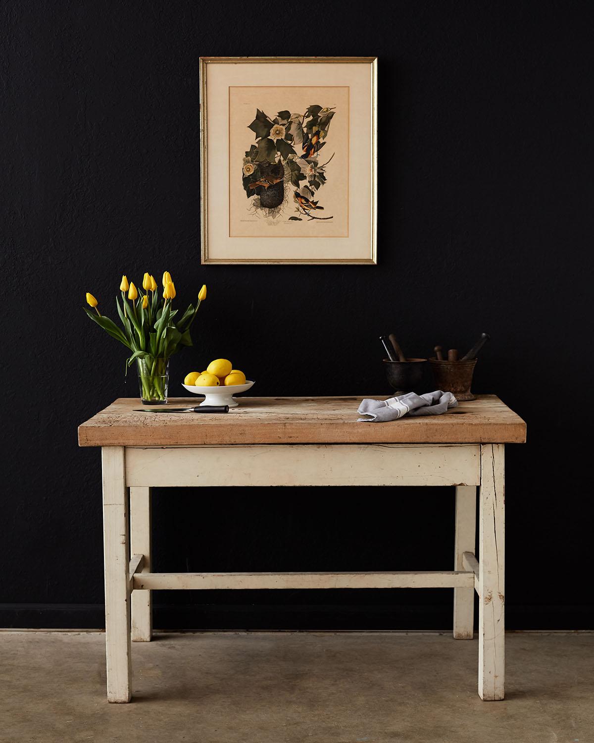 Rustic American farmhouse work table featuring a beautifully worn butcher block top. The top is over 2 inches thick of hardwood, probably maple with a natural light finish. The base is made of square legs and stretchers with a vintage white paint.