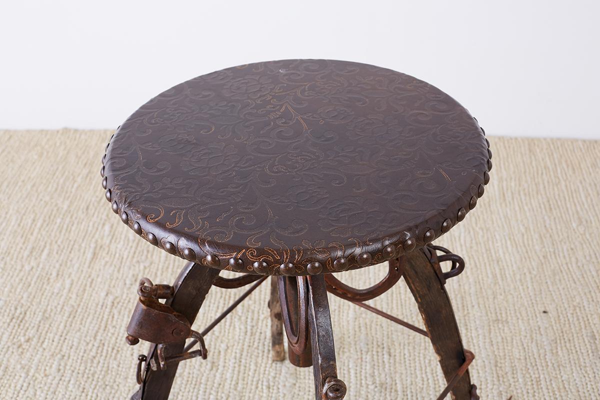 Rustic iron and tooled leather bar stool, or drink table featuring an equestrian motif. Decorated as a saddle or yoke with iron strapping, stirrups, loops, and horseshoes on each leg. Artisan made with a whimsical Americana Folk Art style. The