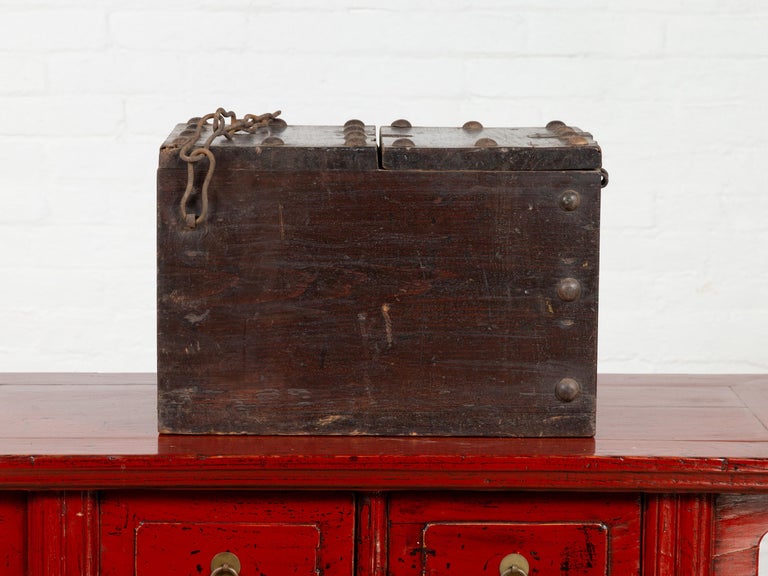 An antique Chinese wooden cash box from the early 20th century, with chain and partially removable top. Charming our eyes with its rustic presence and nicely weathered appearance, this wooden cash box is accented the regular presence of metal studs