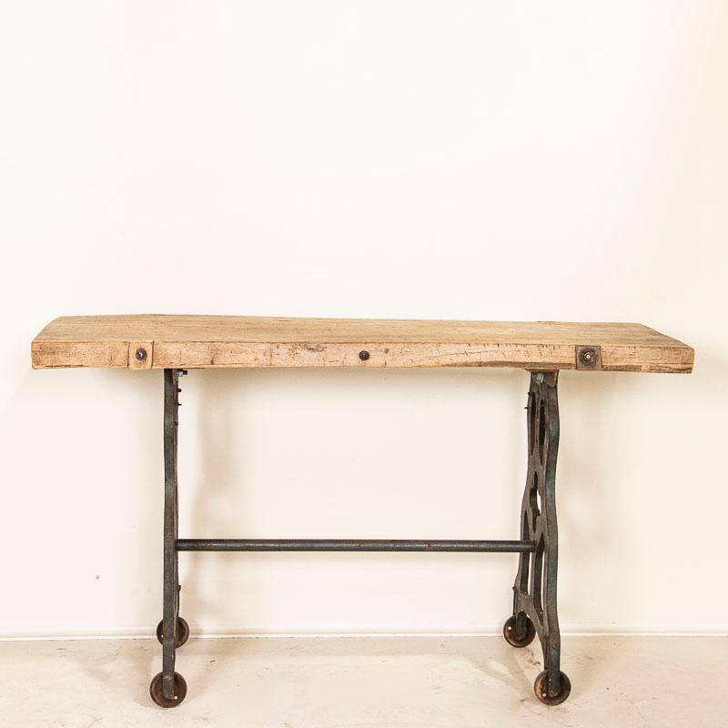 This tall and dramatic console table draws you in to reach out and touch it's thick hard wood slab top that originally served as a work table. The ornate, vintage cast iron legs on castors add stability and give the piece an industrial look in