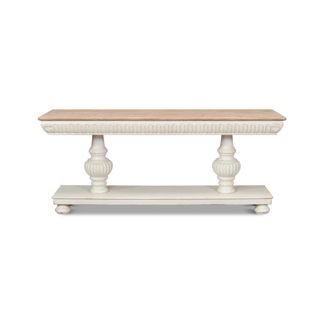 A dramatic transitional pine console, with a natural wooden top above an antiqued white painted base. It features carved volutes around the frieze, atop two carved baluster pedestals. This console table not only serves as a functional surface but