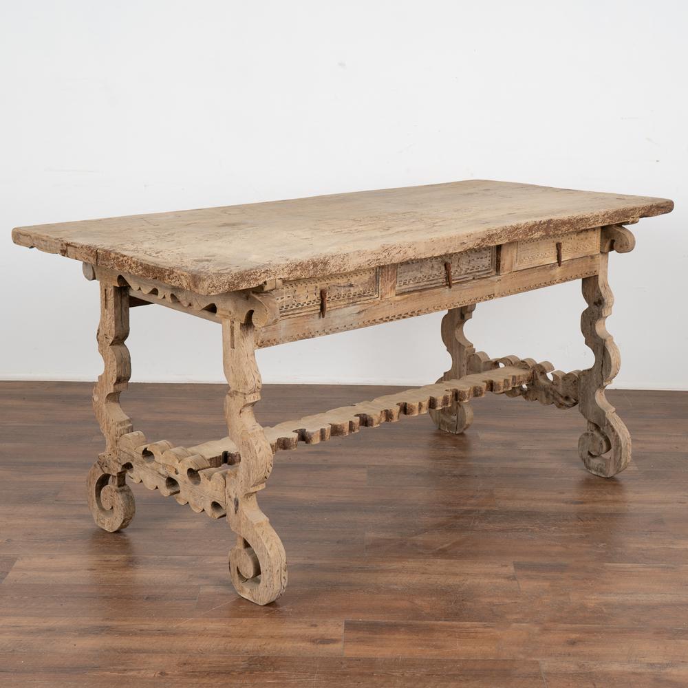 This bleached oak table has strong presence thanks to the elaborate carving that creates the visually impressive harp shaped legs of the trestle base.
Three drawers with rustic pulls on each side allow this to be used a a unique writing table or