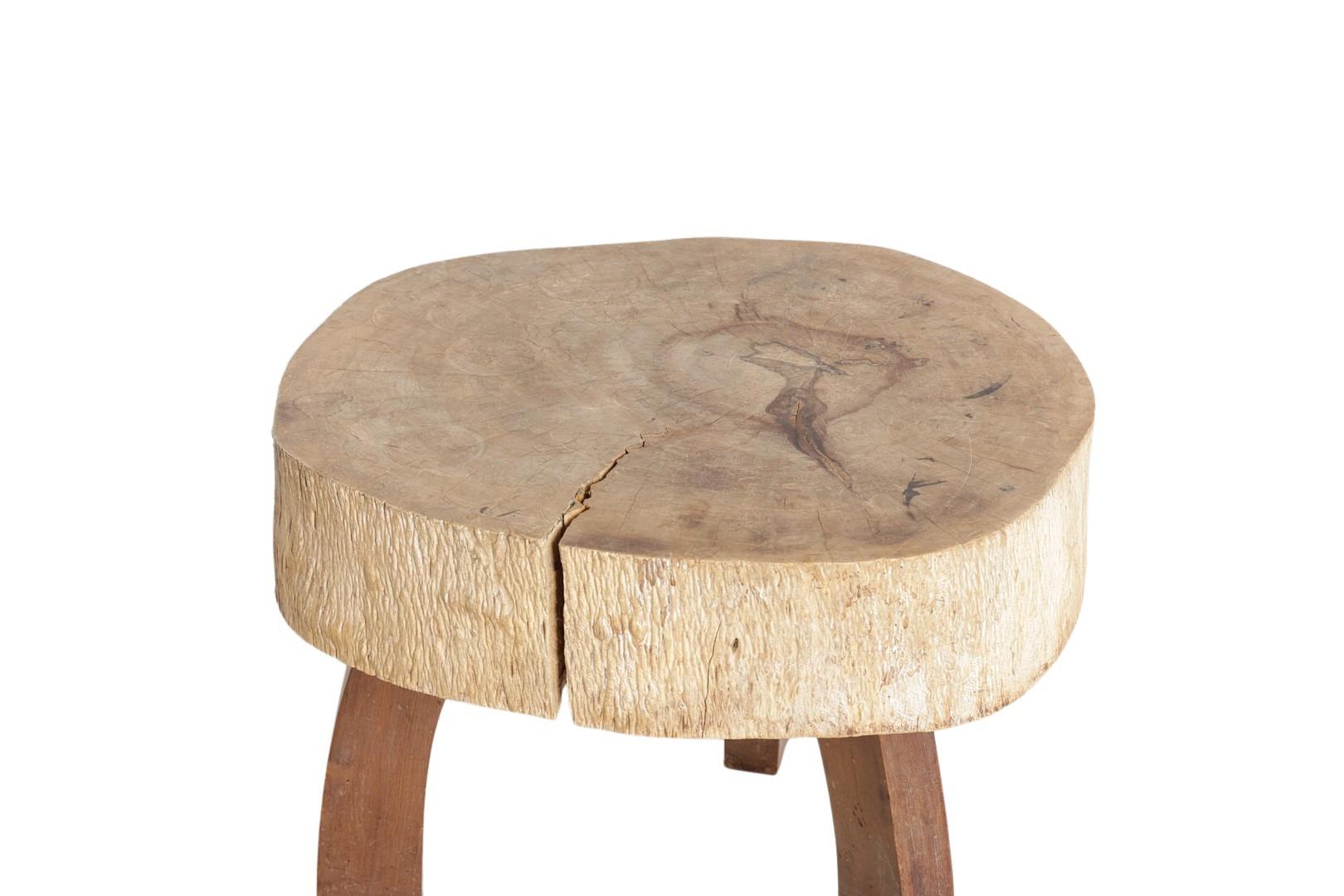 Organic and sculptural in form, this rustic table is crafted from a thick, solid slab of natural wood—most likely suar—set on three gently curved legs. The top features a raw, textured edge and a central split that highlights the wood’s natural
