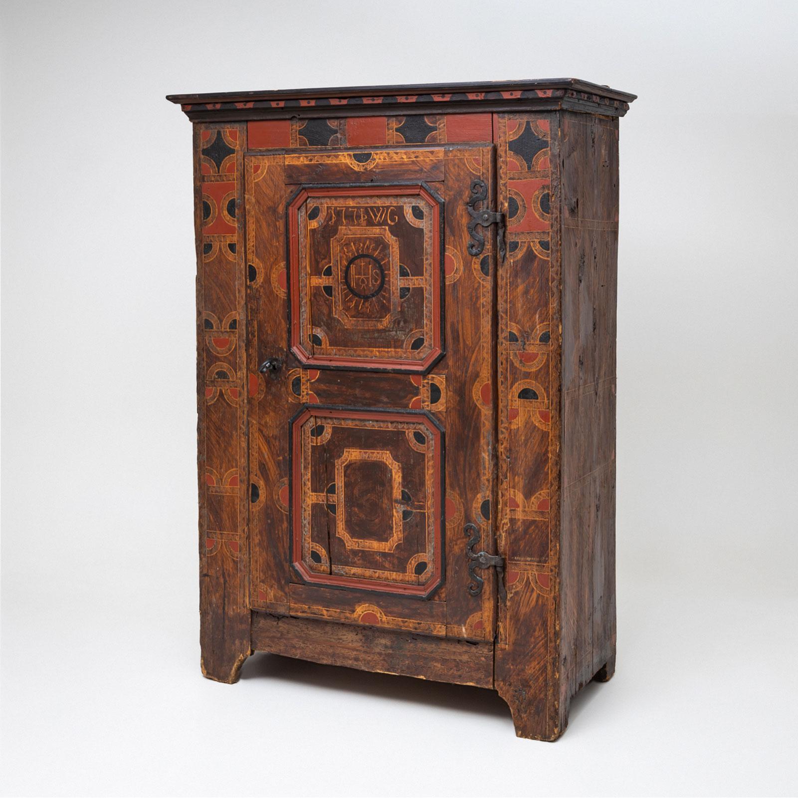 Un mobile rustico a un'anta proveniente dalla regione alpina, realizzato nel 1771. Il mobile poggia su piedi a staffa sagomati e presenta una finitura verniciata in rosso e nero che ne evidenzia il carattere tradizionale.

La porta è ornata da