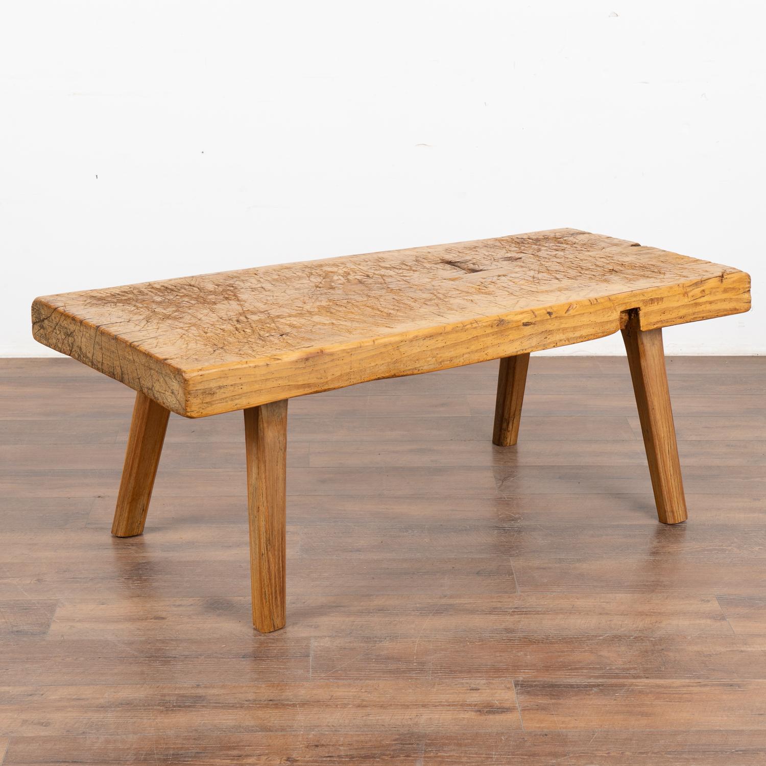 The appeal of this rustic coffee table comes from the thick hard wood top that originally served as a work table, resting on strong squared peg splay legs. 
Note the heavy gouges, nicks, old cracks and stains that all combine to build the vintage