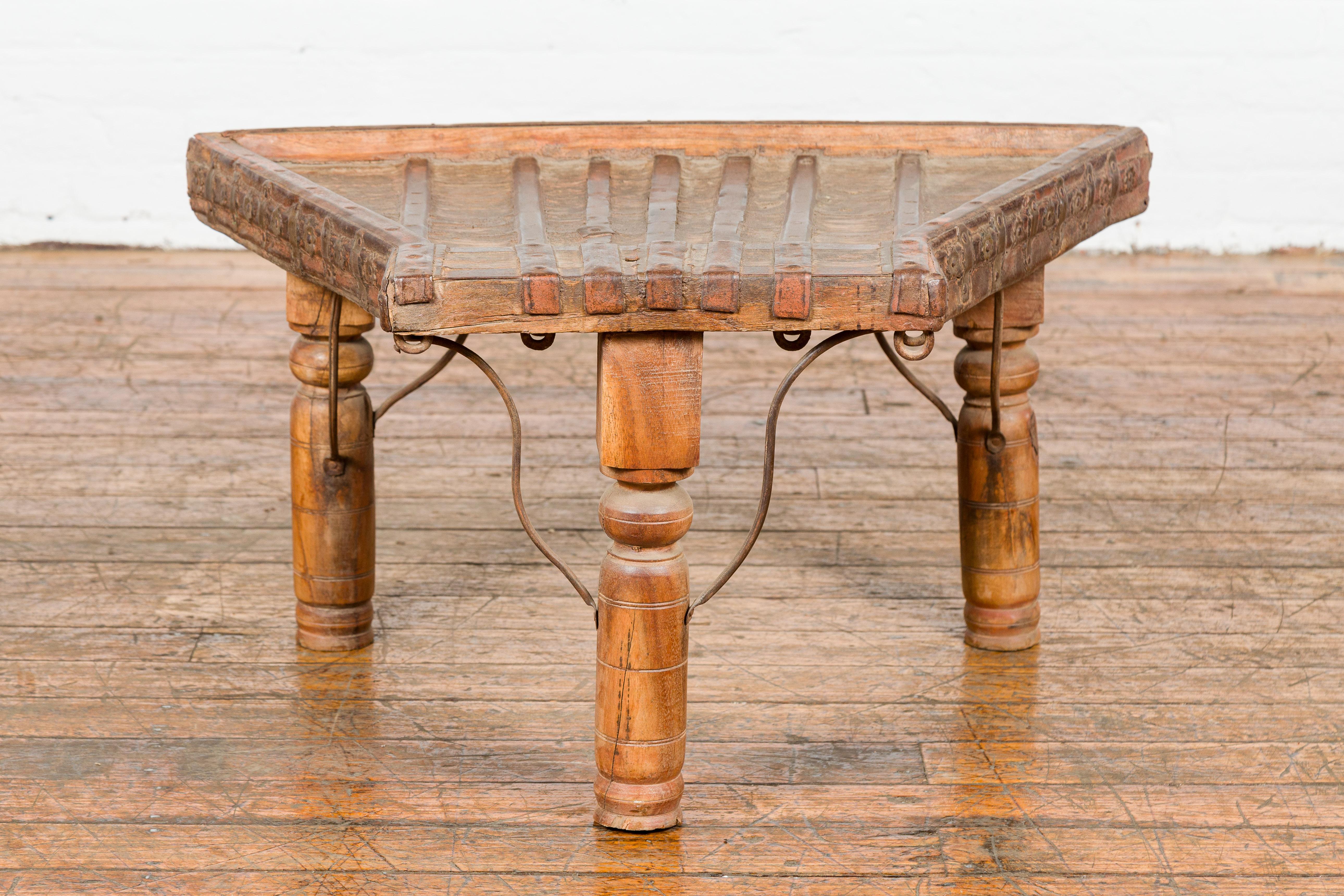An antique rustic Indian handmade coffee table from the 19th century with trapezoidal top, protruding front, curving iron stretchers, floral shaped studs on the apron, turned baluster legs and nicely weathered patina. Behold the rustic charm of this