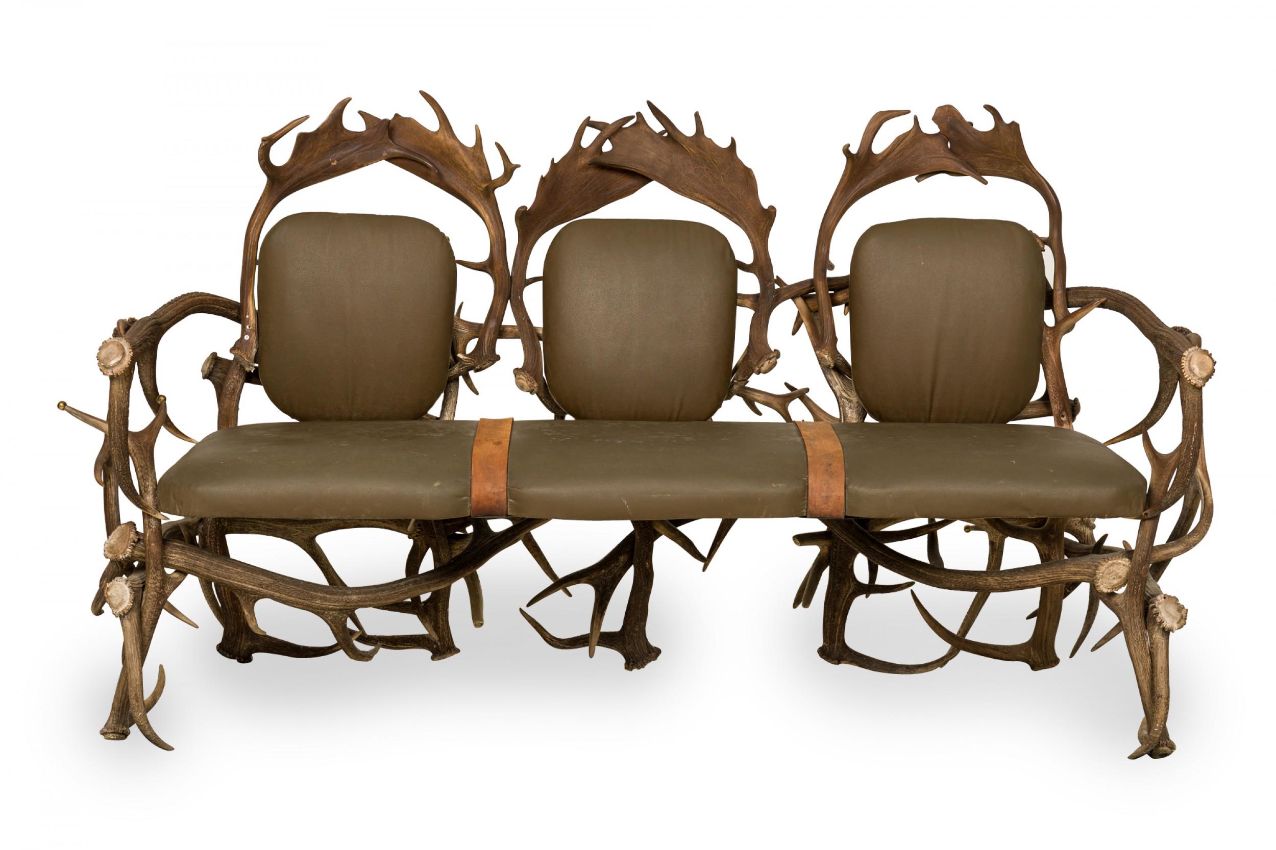 Rustic Continental-style three seat settee with dark olive green leather upholstered seat and backs with two decorative brown suede straps in a frame built from resin faux moose and deer antlers, some of which are capped with decorative brass balls.