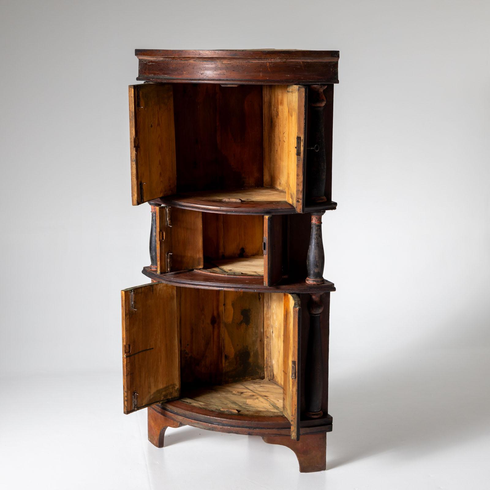 Rustic corner cupboard with baluster columns and three-part design. The cabinet stands on cut-out feet and is painted dark red. The columns are painted a contrasting black colour. 