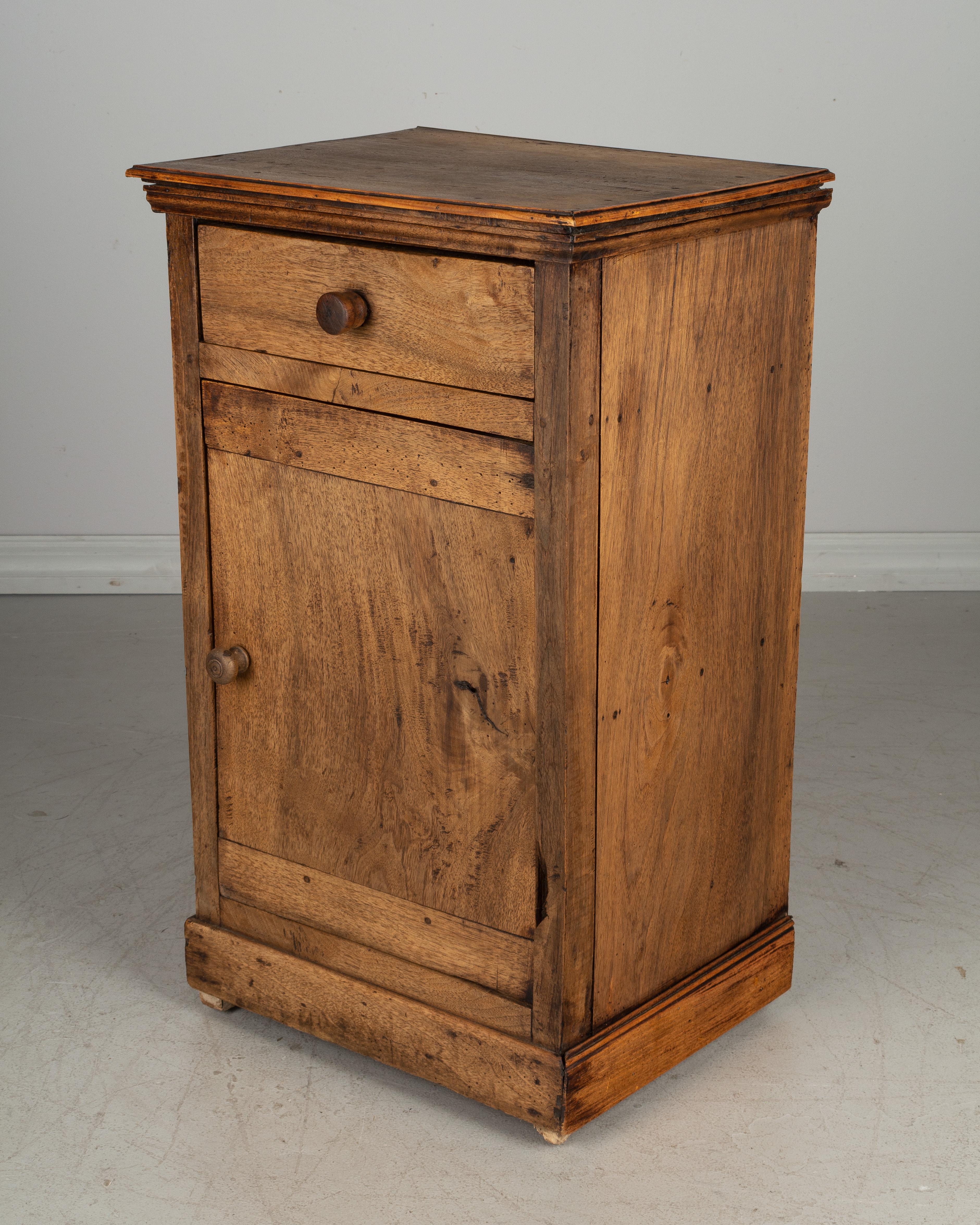 A rustic Country French side table or nightstand made of thick planks of solid walnut with faded distressed finish. Small dovetailed drawer above a cabinet door opening to one shelf. Large turned wood knobs. Please refer to photos for more details.