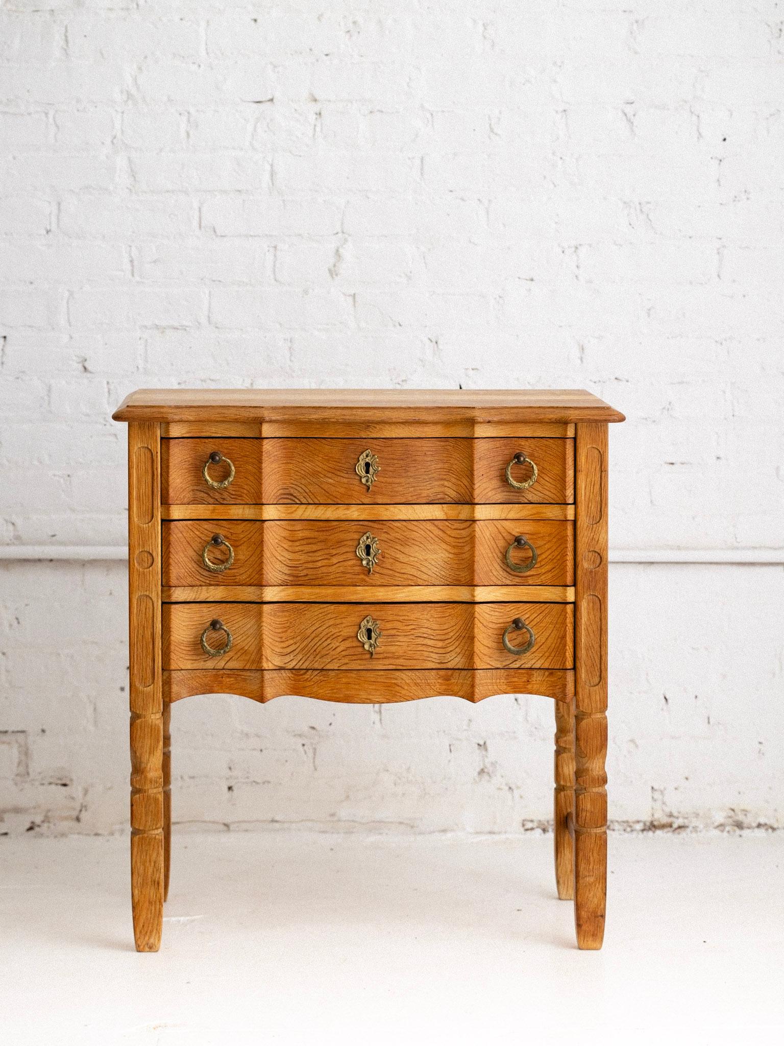 A rustic Danish modern small cabinet or nightstand in the style of Henning Kjaernulf. Three drawers with brass handles and serpentine silhouette. No key present.