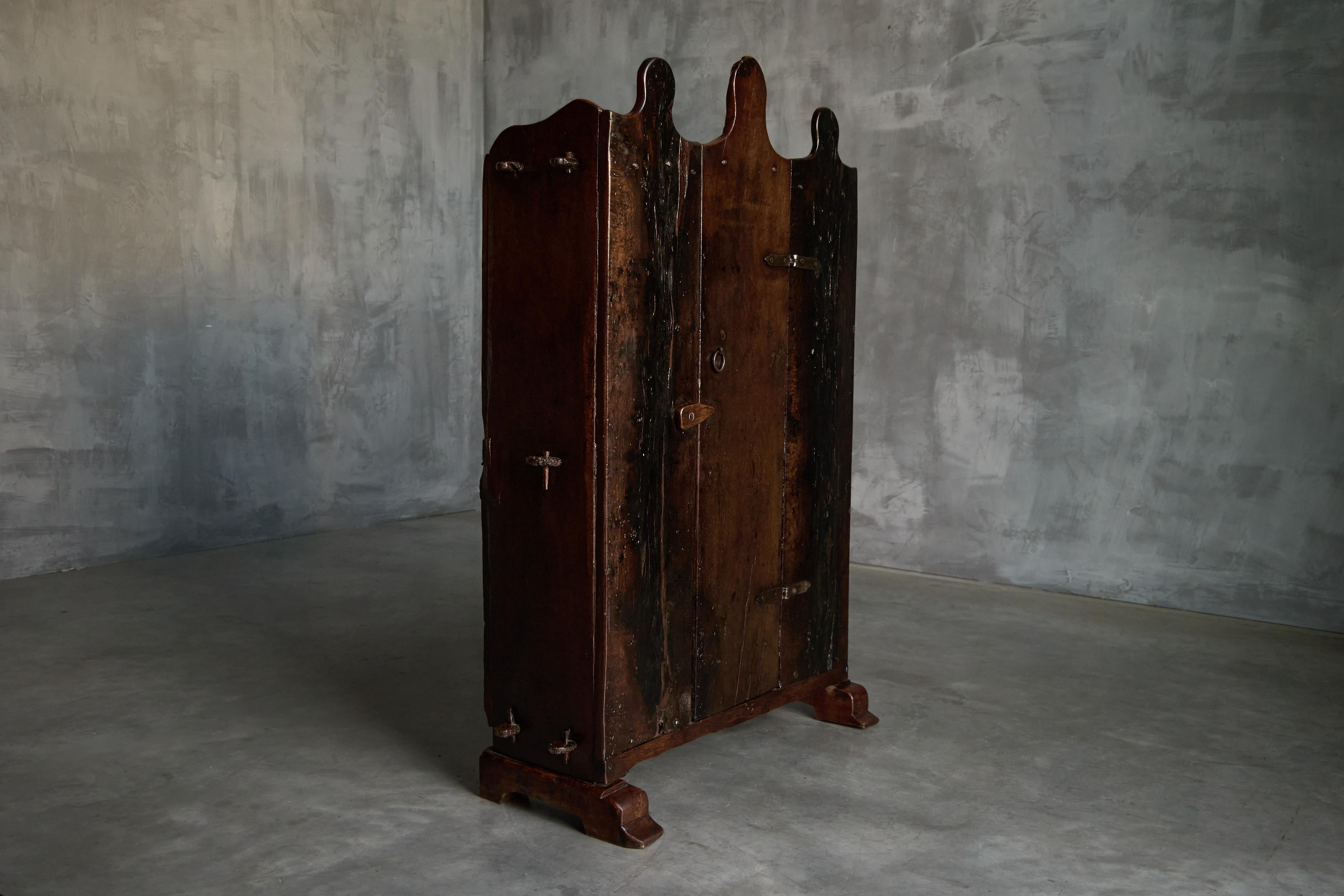 Cette charmante armoire rustique provenant de l'Espagne du XIXe siècle est fabriquée en bois massif foncé et présente une belle patine d'usage. Son design se caractérise par un sommet festonné distinctif, qui ajoute une touche d'élégance à son