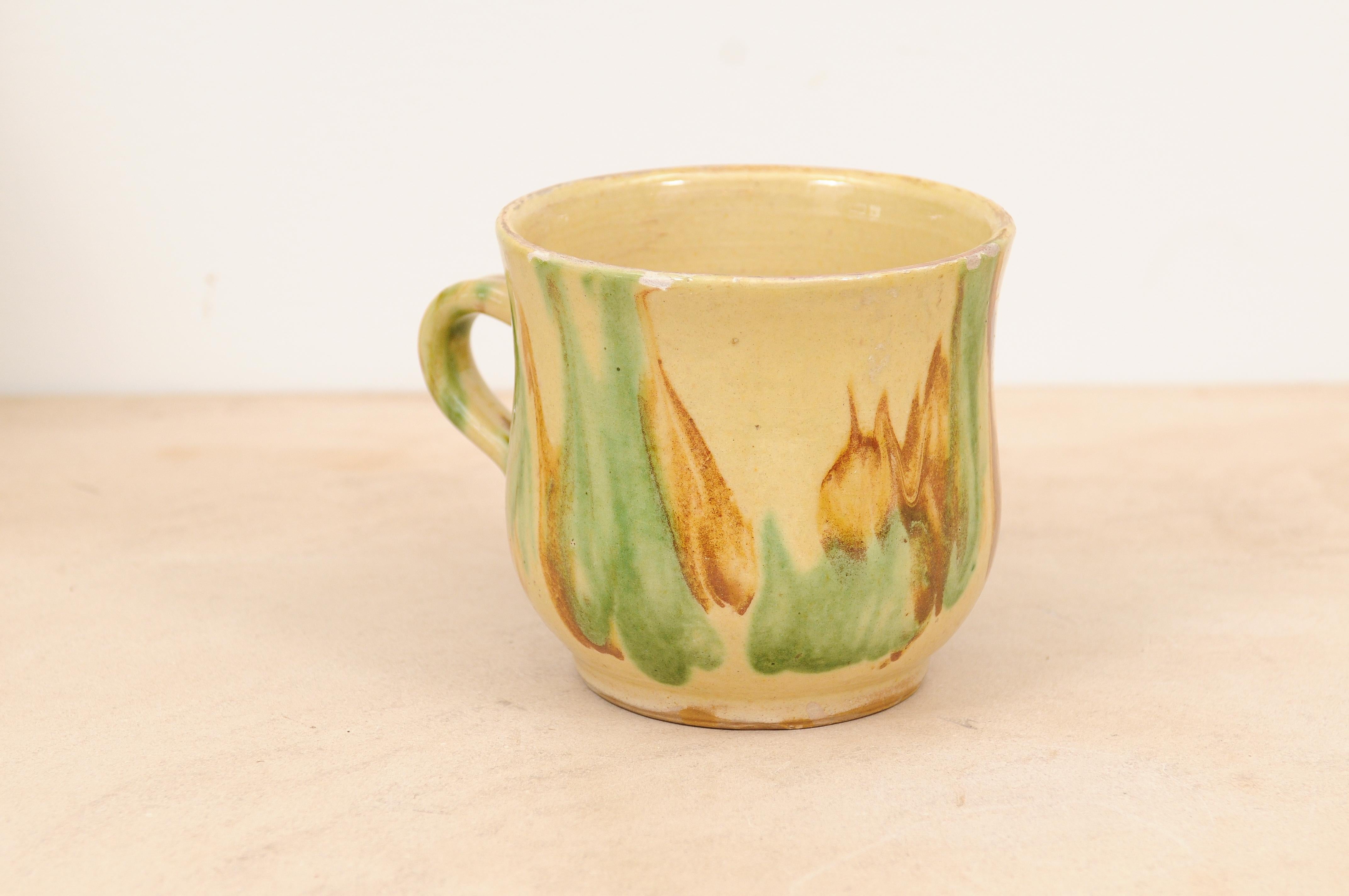 A French pottery mug from the 19th century, with yellow, green and rust glaze. Created in France during the 19th century, this pottery mug features a cylindrical curving body showcasing a yellow ground adorned with green and rust glaze. Presenting a