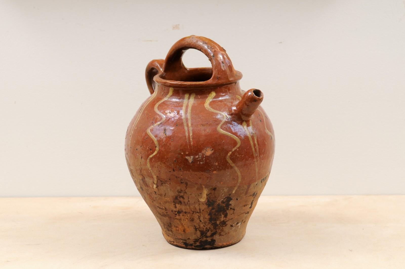 A rustic French glazed terracotta oil jug from the 19th century, with wavy lines and nicely distressed appearance. Created in France during the 19th century, this glazed terracotta pottery piece features a brown circular body accented with golden