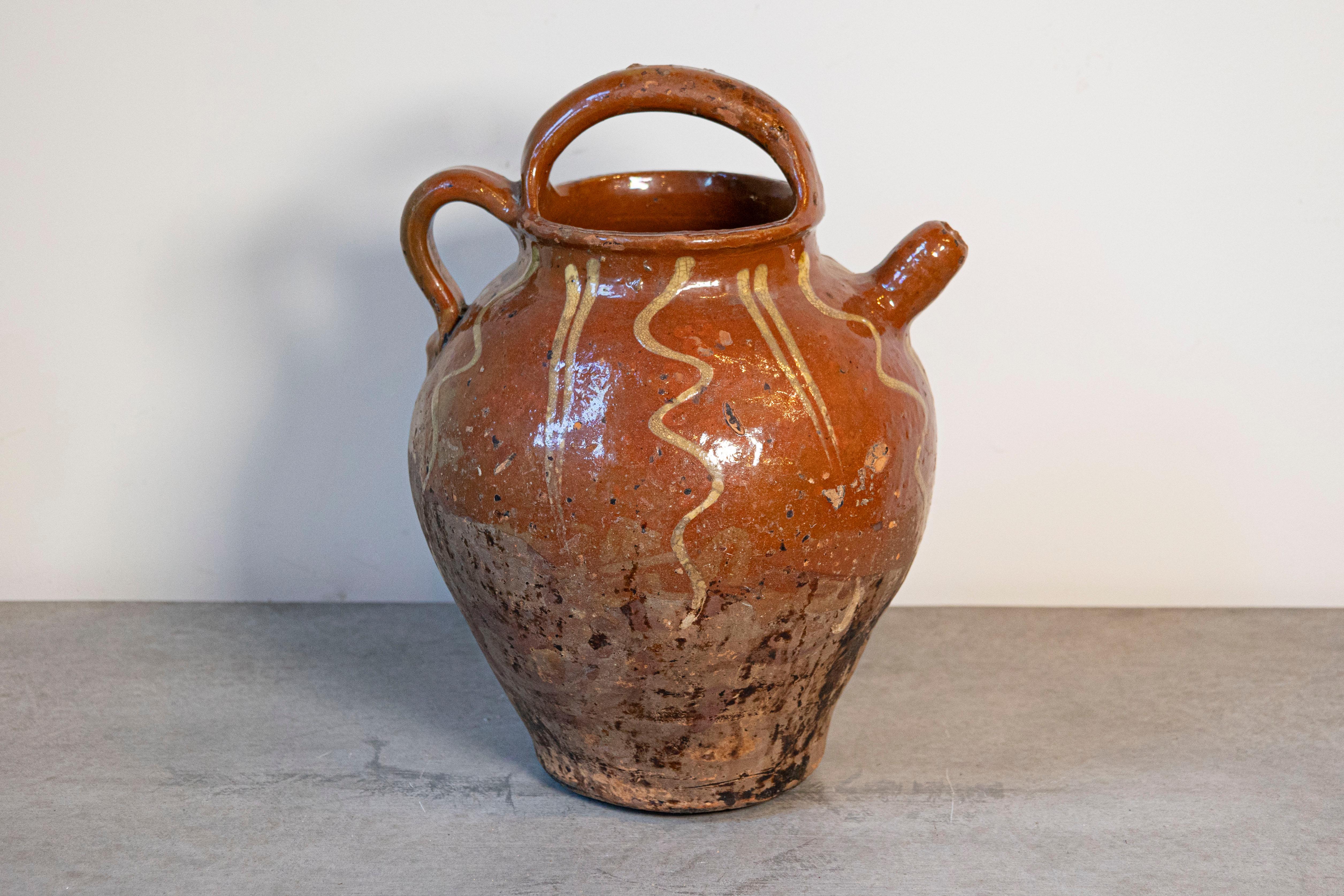 A rustic French glazed terracotta oil jug from the 19th century, with wavy lines and nicely distressed appearance. Created in France during the 19th century, this glazed terracotta pottery piece features a brown circular body accented with golden