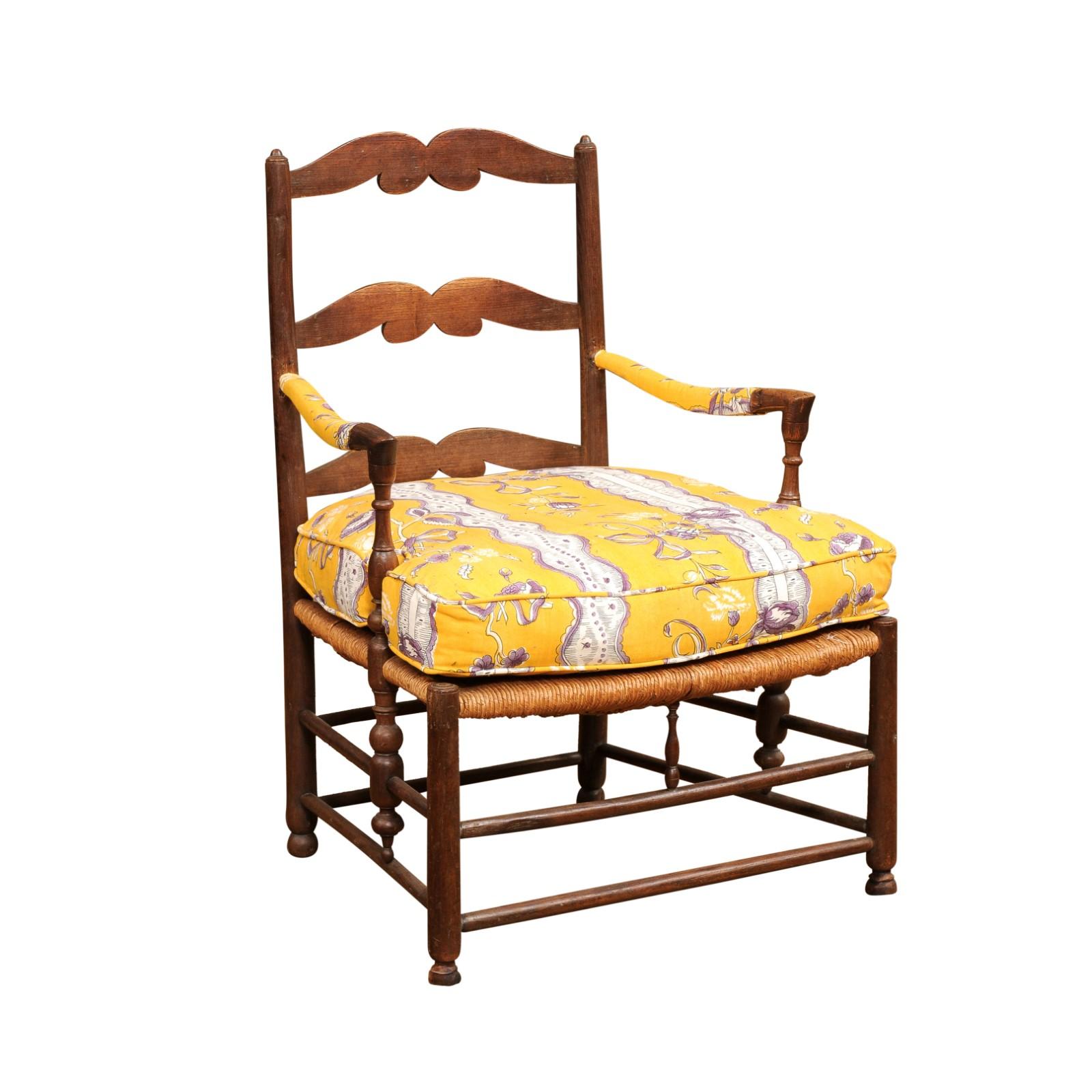 A rustic French walnut armchair from the late 18th century with rush seat, carved ladder back, curving arms and upholstered cushion. Created in France during the later years of the 18th century, this walnut armchair charms us with its rustic