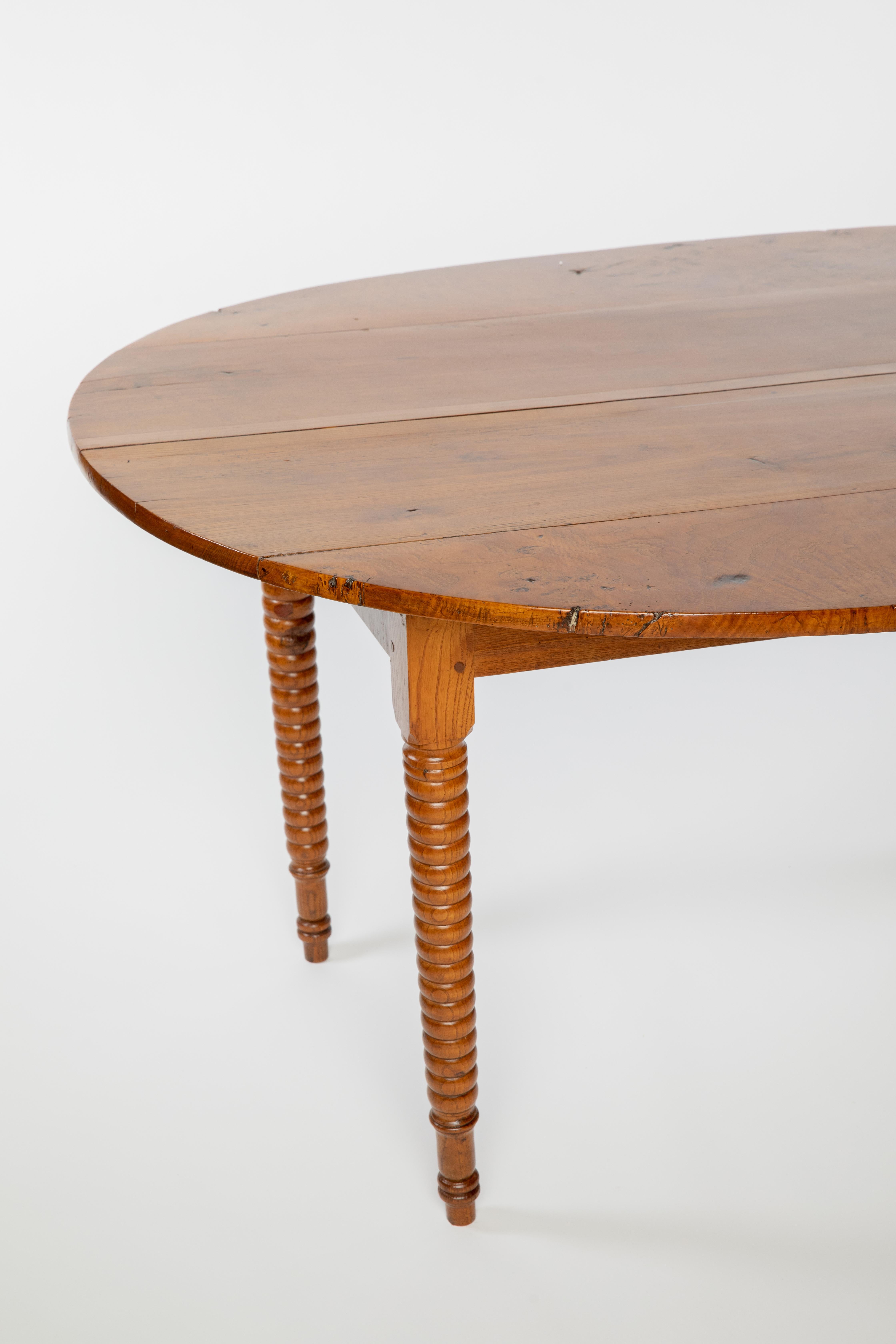 Rustic French mixed wood farmhouse table, 19th c., having oval-shaped plank top, over spool-turned legs, ending in blunt arrow feet.