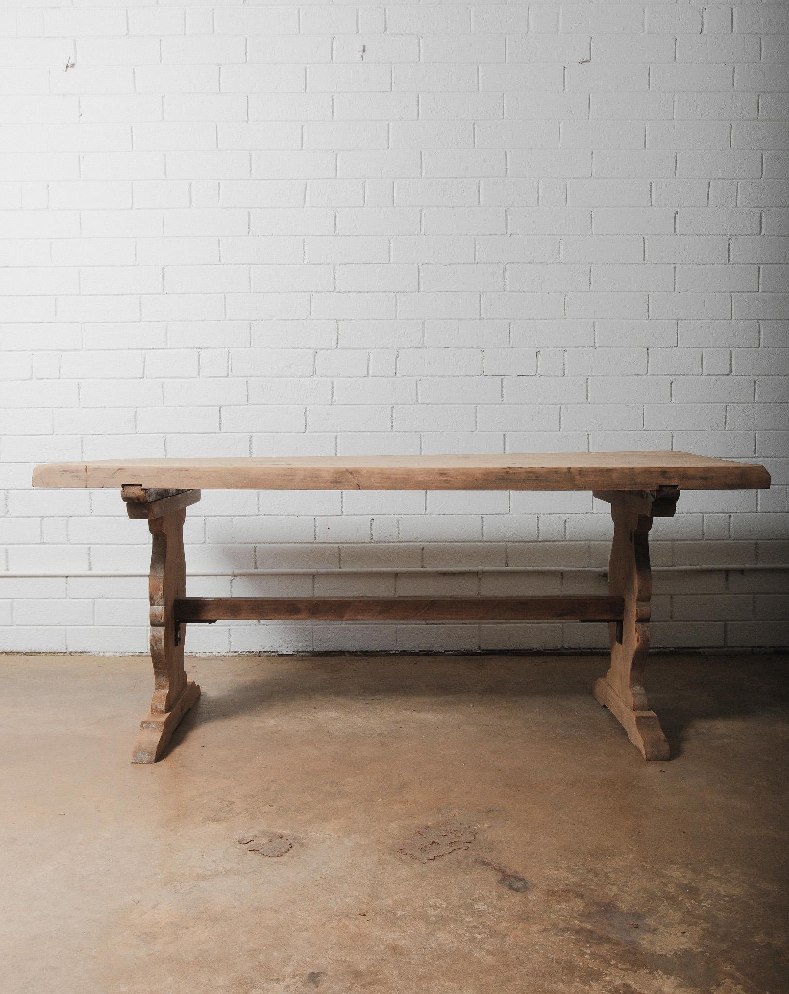 This rustic trestle dining table features a solid plank top with smoothed edges and a generously thick profile. The carved pedestal bases are connected with a single stretcher and anchored by wedged joinery, providing both architectural interest and