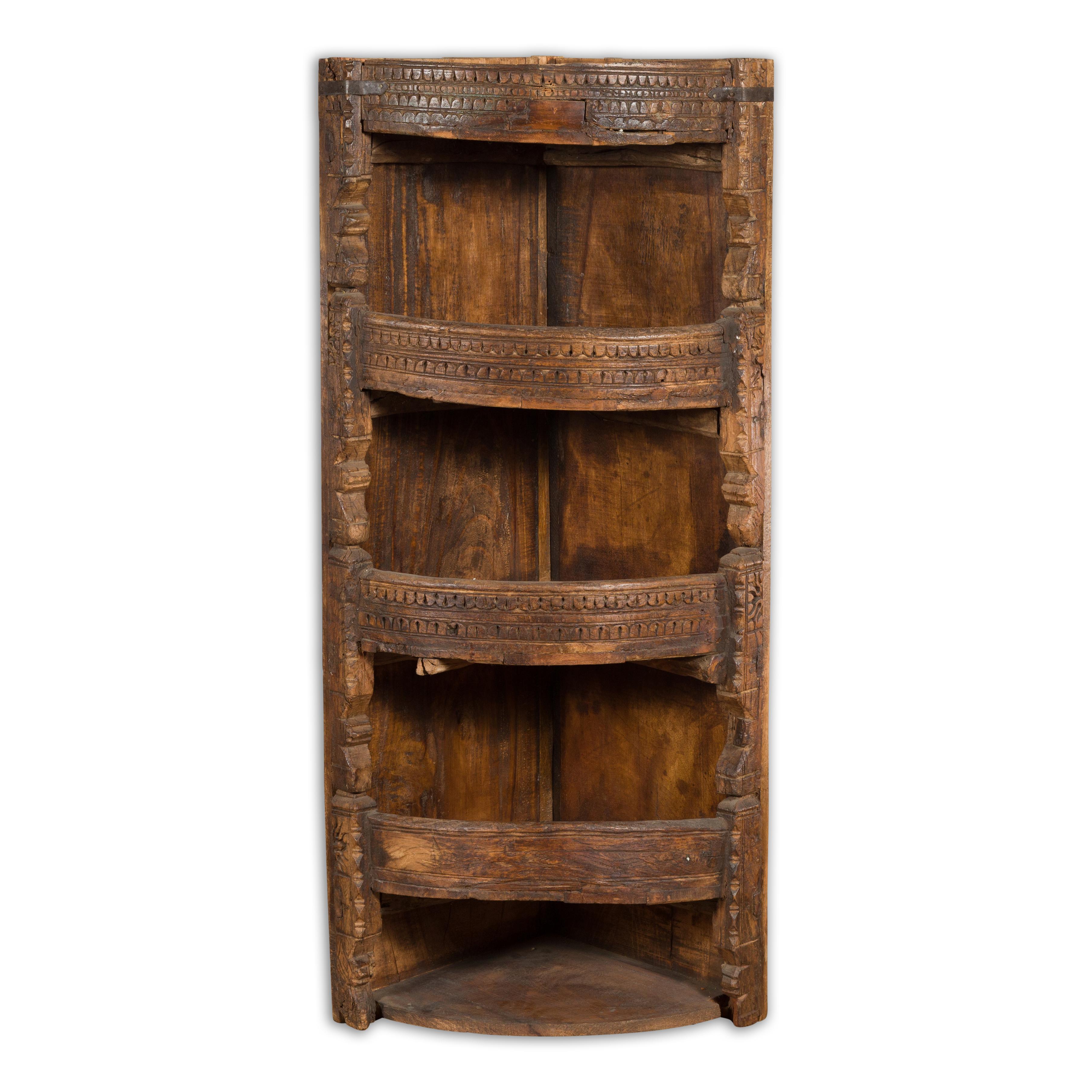 A small Indian rustic wooden corner display cabinet from the 19th century with carved geometric motifs, bow front shelves and weathered appearance. Created in India during the 19th century, this antique wooden corner cabinet charms us with its small