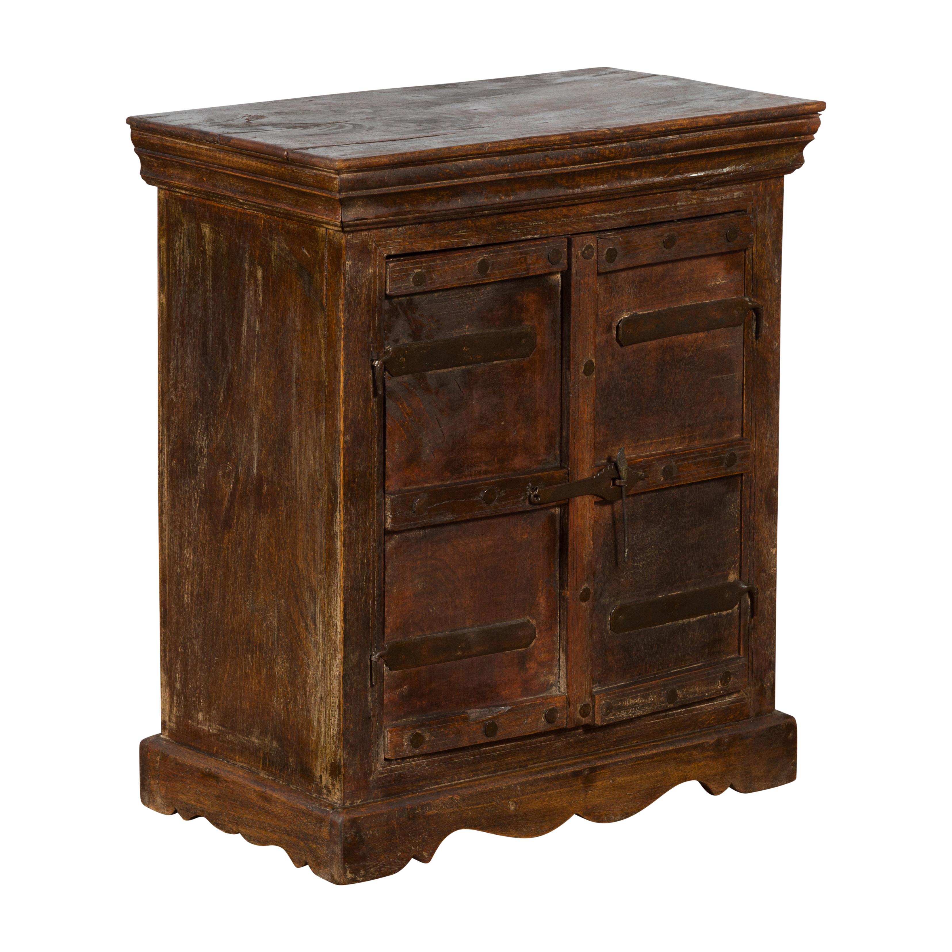 A vintage Indian sheesham wood bedside cabinet from the mid 20th century with dark finish, iron hardware, scalloped plinth and rustic character. Created in India during the Midcentury period, this rustic small sheesham wood bedside cabinet features