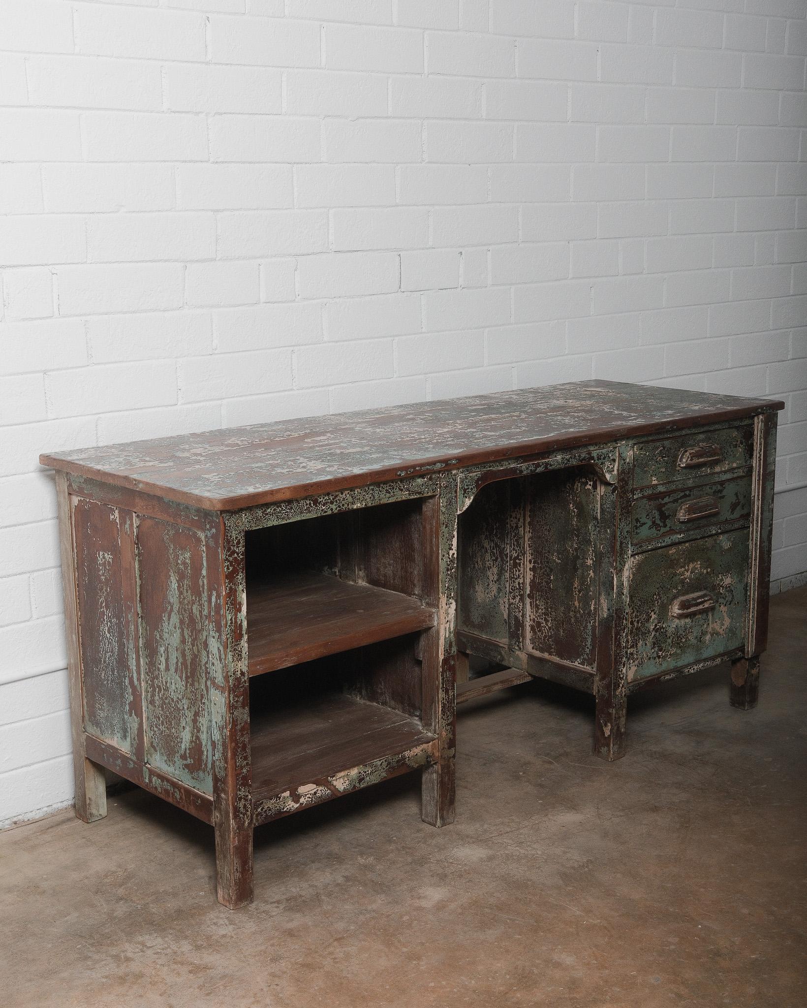 This vintage wooden desk exudes rustic industrial charm with its weathered green patina and distressed finish. Crafted from solid wood, the desk features a spacious work surface, open shelving on one side, and three deep drawers with sturdy metal
