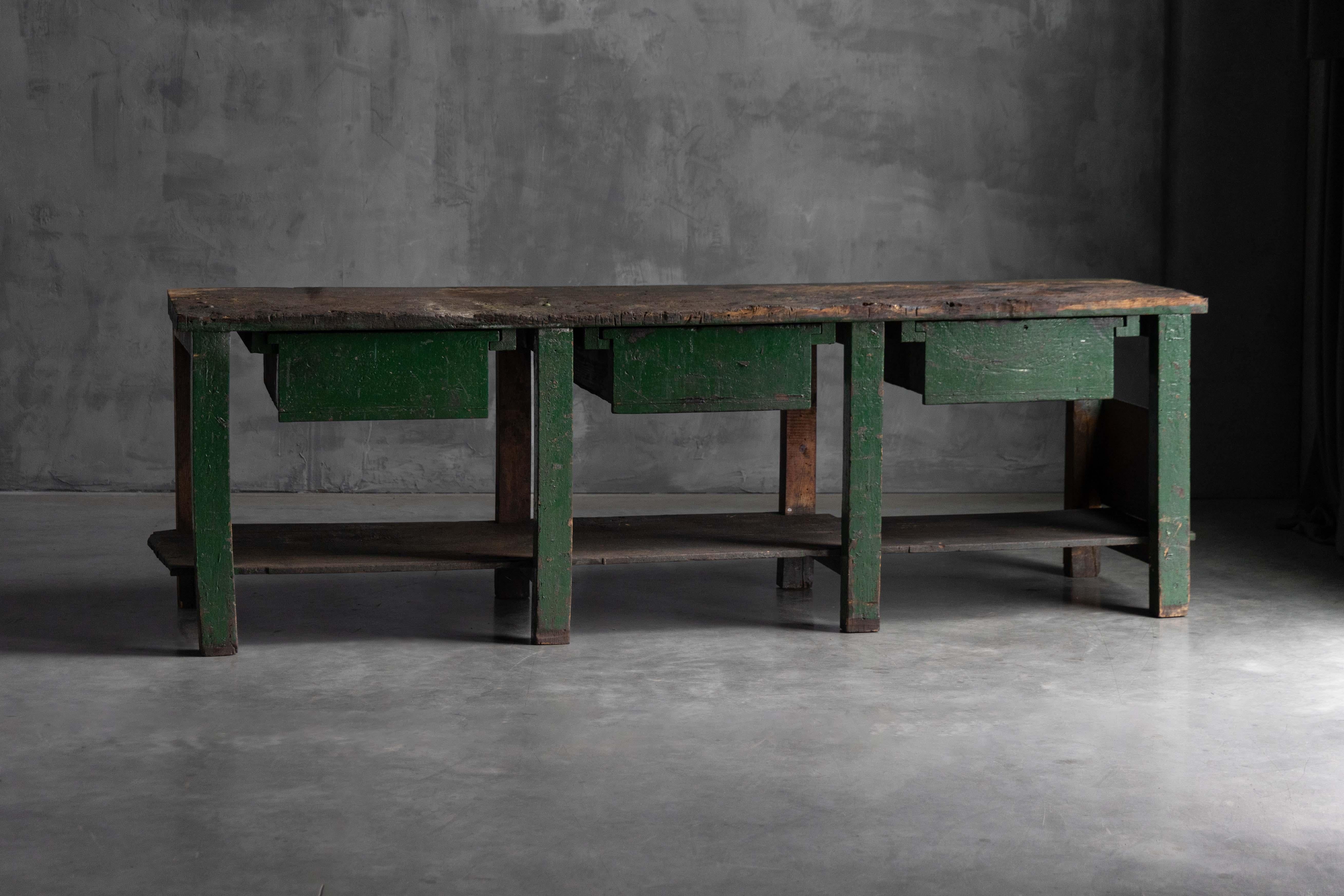 This early 20th-century rustic industrial workbench, with its deep green painted base and thick, timeworn wooden top, carries the rich patina and character of decades of practical use. Three spacious drawers with simple dividers and a full-length