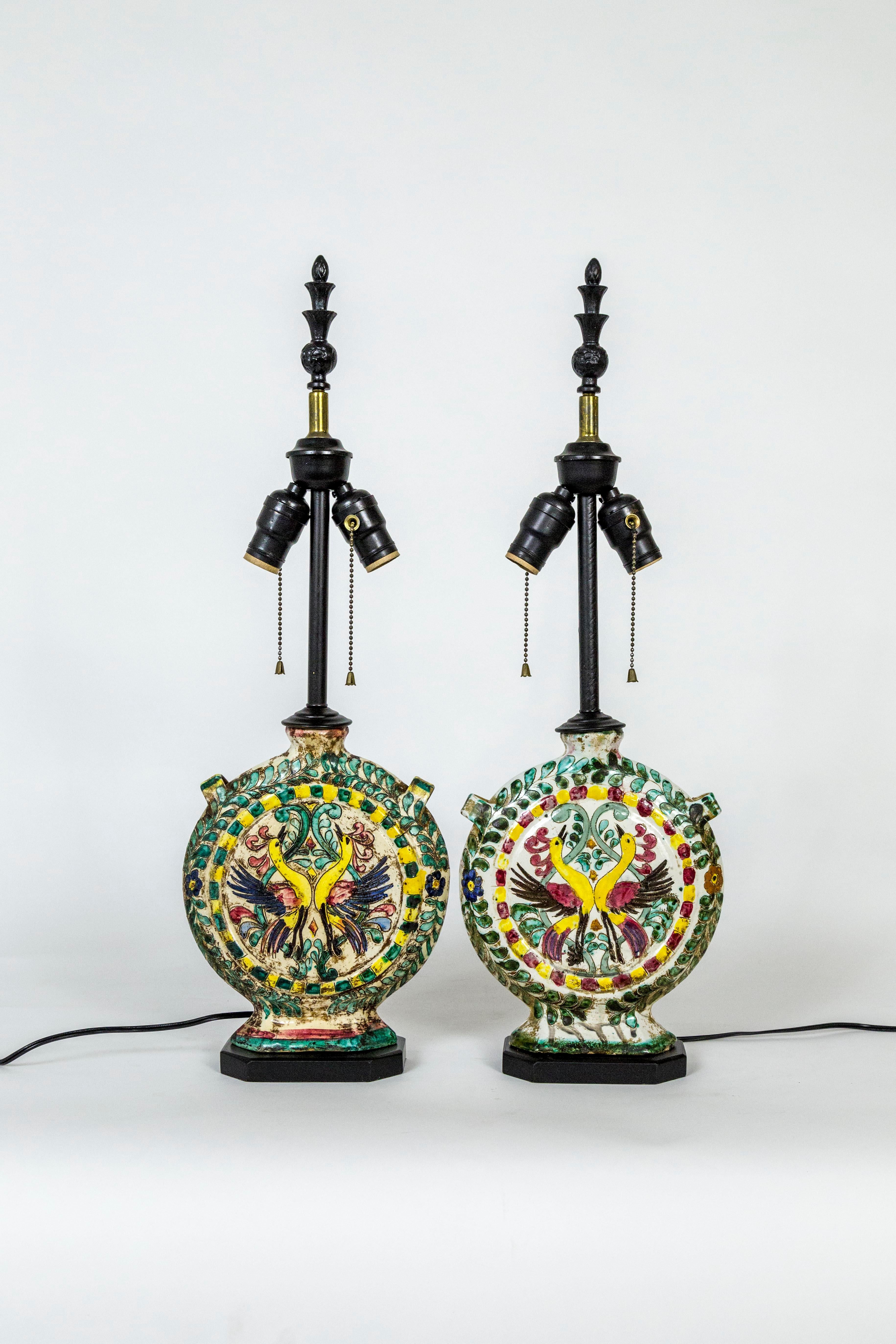 Narrow, ceramic wine canteens with painted birds, primarily yellow, green, and white in color, as rustic table lamps. Italian, Majolica tradition, Folk Art, circa 1900. On black, wood base, with black, metal finials; 2 sockets. Newly wired.