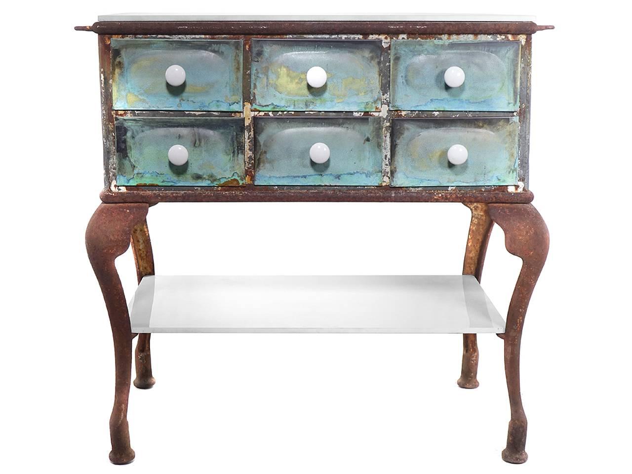 This is a bow leg medical work table with an amazing patina. In this case the patina makes a bigger statement the design of the table itself. The top and glass shelf are heavy milk glass as are the draw pulls.