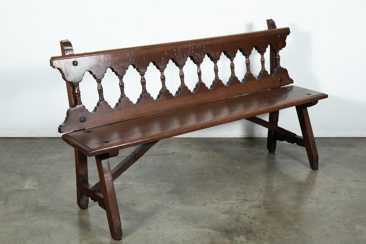 Rustic mid-19th century Spanish bench handcrafted of walnut in the Catalan region of Spain, having a carved back rail over a gallery of turned spindles, large iron nail heads, and a plank seat, circa 1850s. Raised on a trestle base joined by walnut