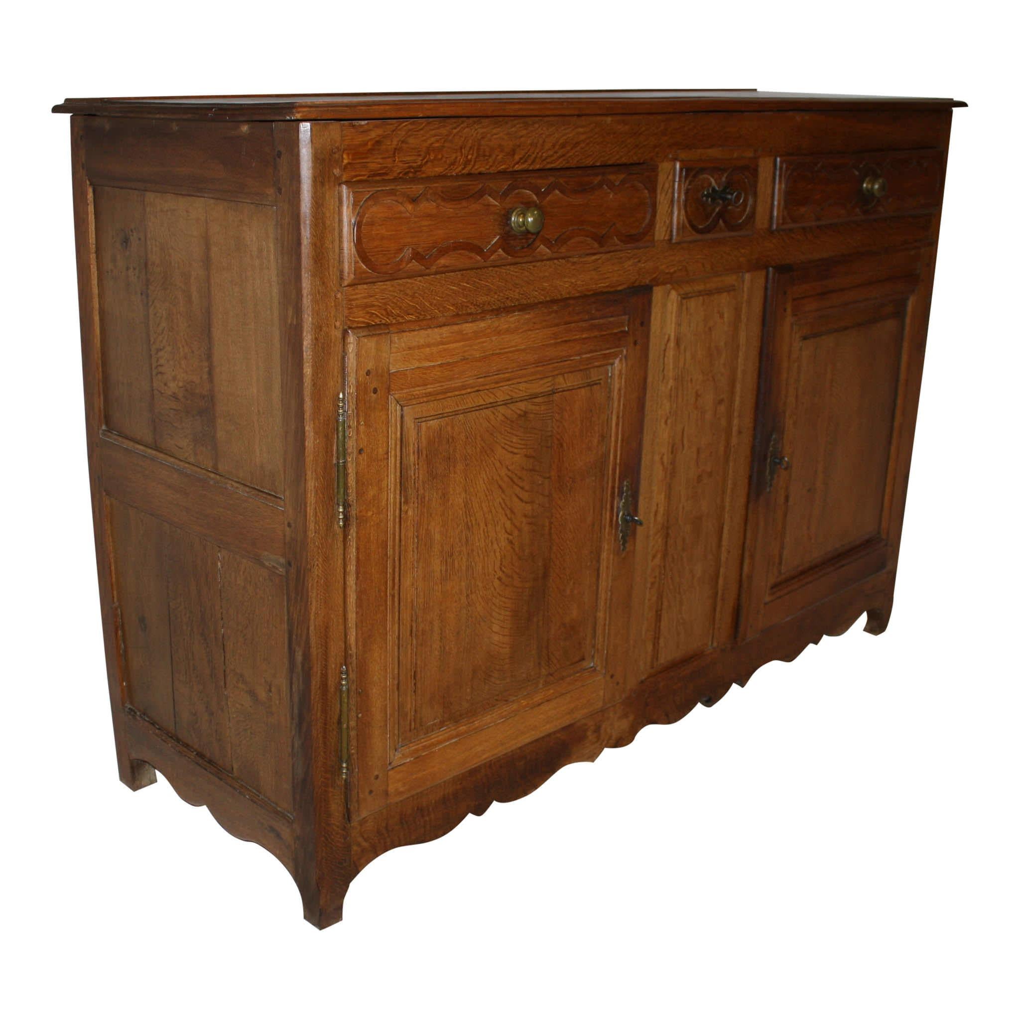 This rustic, oak sideboard, which was crafted in the late 19th century, features a beveled top with a plate groove; a narrow center drawer flanked by wider side drawers with brass knobs; double, raised panel doors with a raised, center panel between