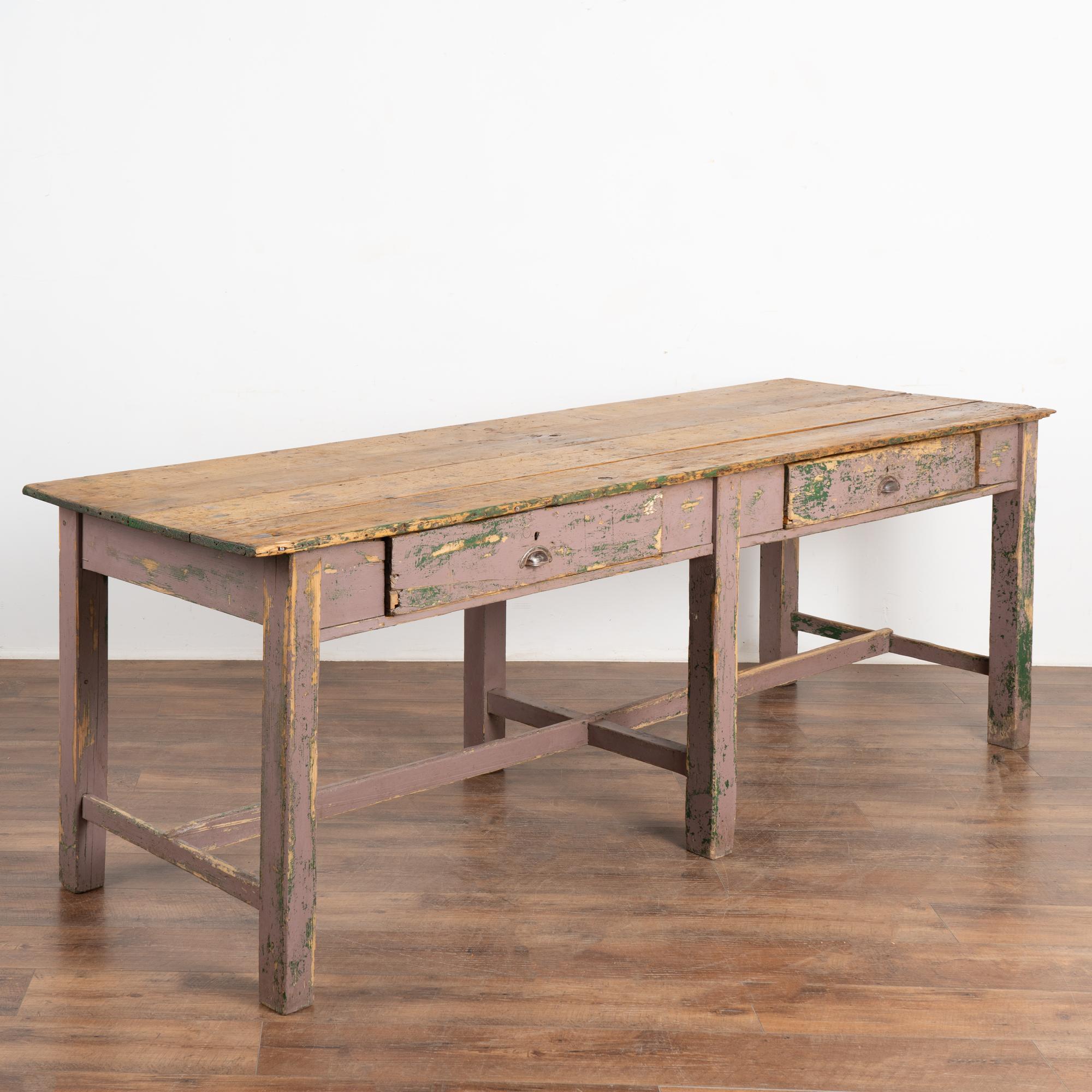 Rustic work table with two drawers and lower stretcher base with six legs. Original purple and green painted finish show extensive distress and wear down to the natural pine below. 
This heavily used table reflects generations of use in every gouge,