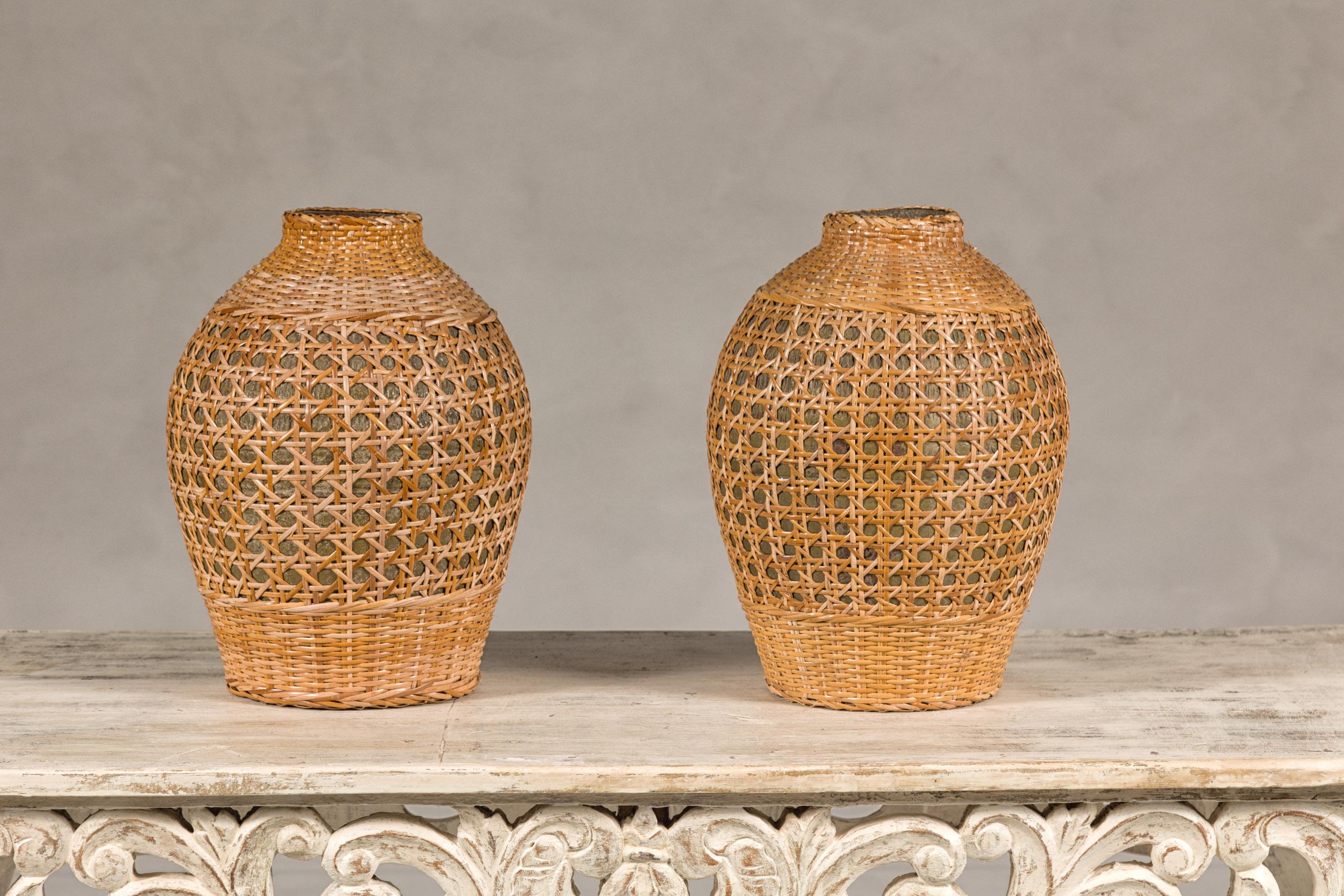 A pair of Midcentury cane over ceramic wicker vases with rustic character. This pair of Midcentury vases embodies a unique fusion of materials, where the traditional craft of cane weaving meets the timeless elegance of ceramic. Originating from