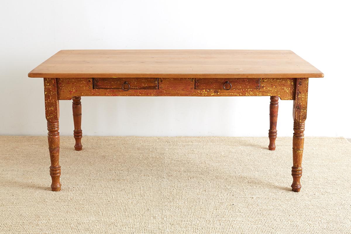 Quintessential rustic pine farmhouse table with old lacquer paint remnants scraped down. Features a 1 inch thick plank top supported by four large turned legs. The table has two drawers on one side for storage with old iron ring pulls. The piece is