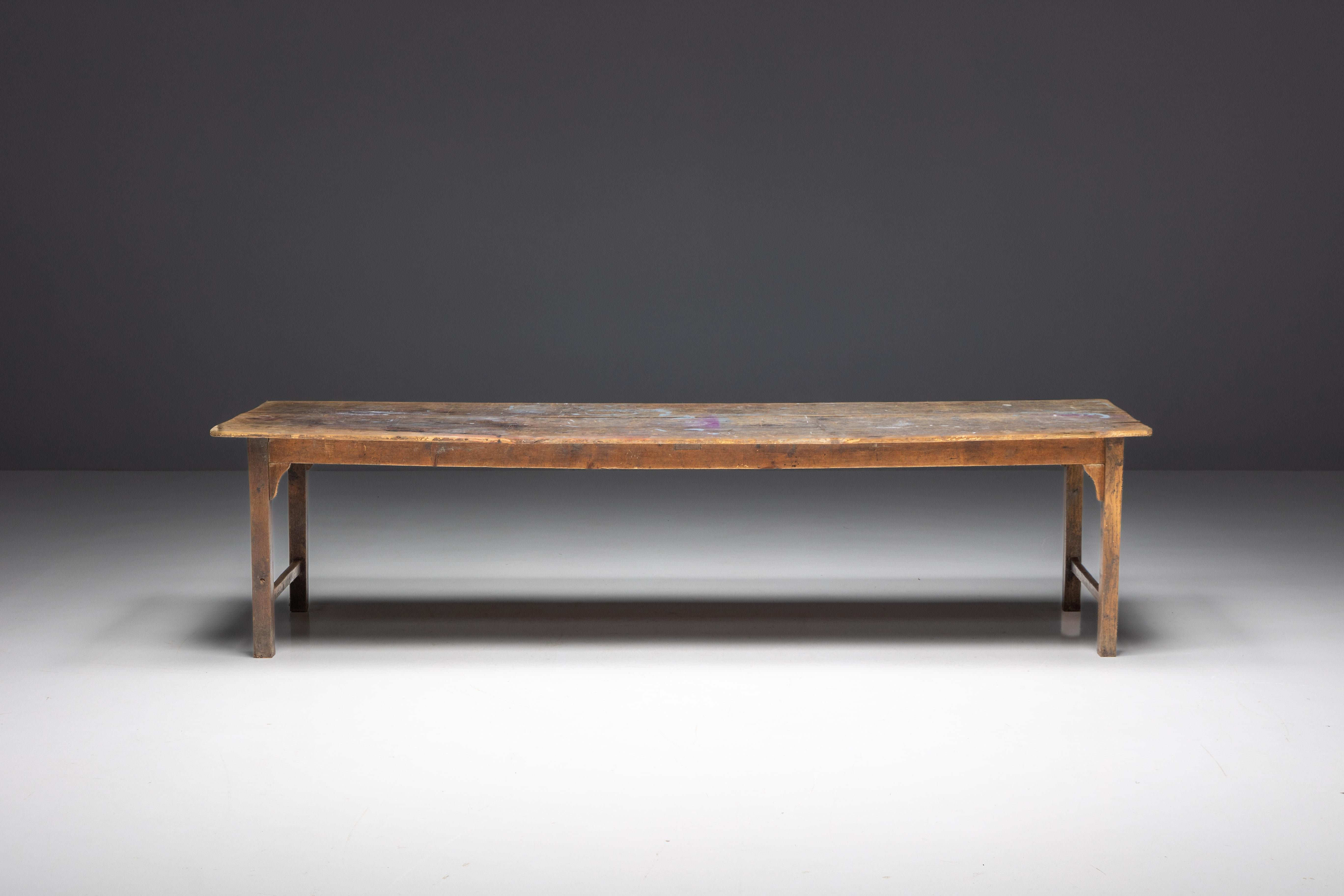 Table de ferme française du XIXe siècle à la patine naturellement vieillie, incarnant la beauté intemporelle de la campagne française. La surface de la table, ornée d'éclaboussures de peinture colorées provenant d'activités créatives passées, ajoute
