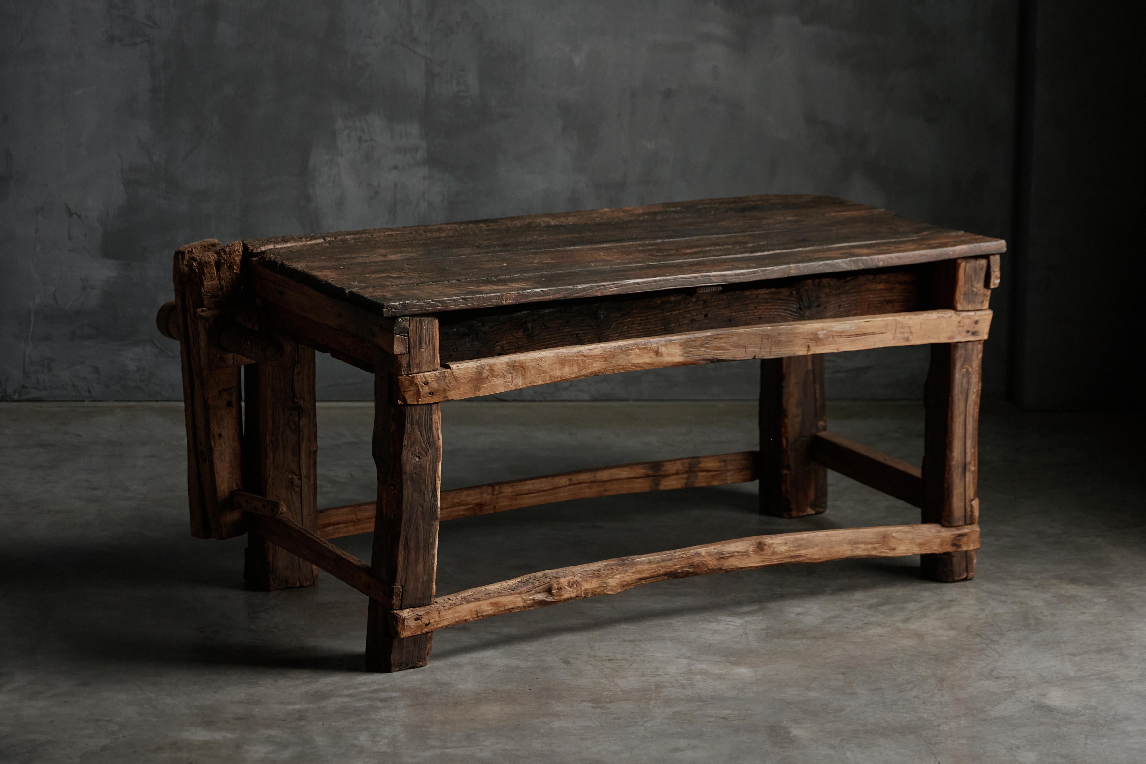 Rustic working bench that exudes a raw, handcrafted charm, showcasing the wear and patina from decades, if not centuries, of use. Made from solid, heavy wood, the bench features a sturdy rectangular tabletop, weathered and worn with deep grain