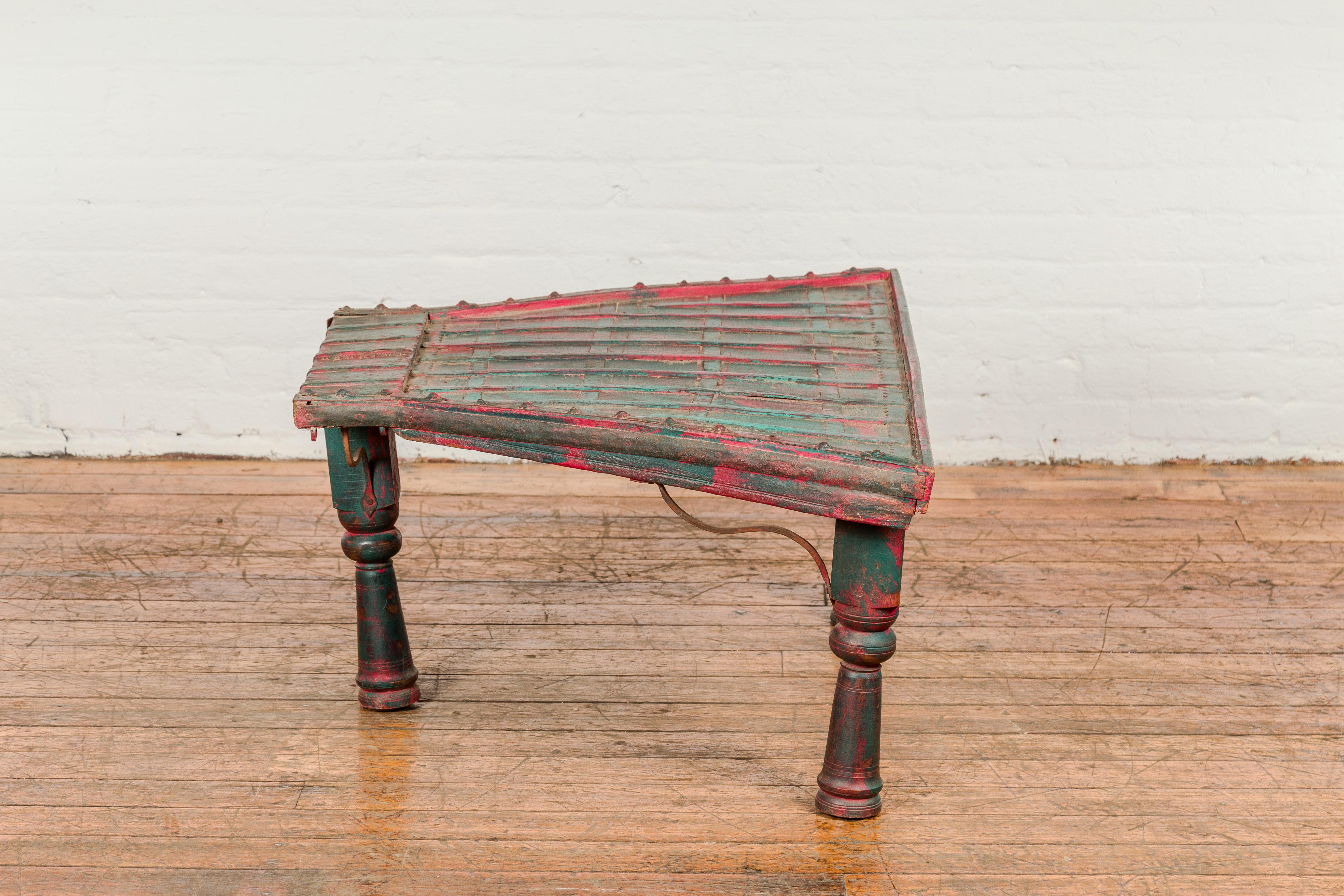An antique rustic green and red painted coffee table crafted from a repurposed bullock cart from the 19th century with trapezoidal top, protruding front, brass floral studs, curving iron stretchers, turned baluster legs and nicely weathered patina. 