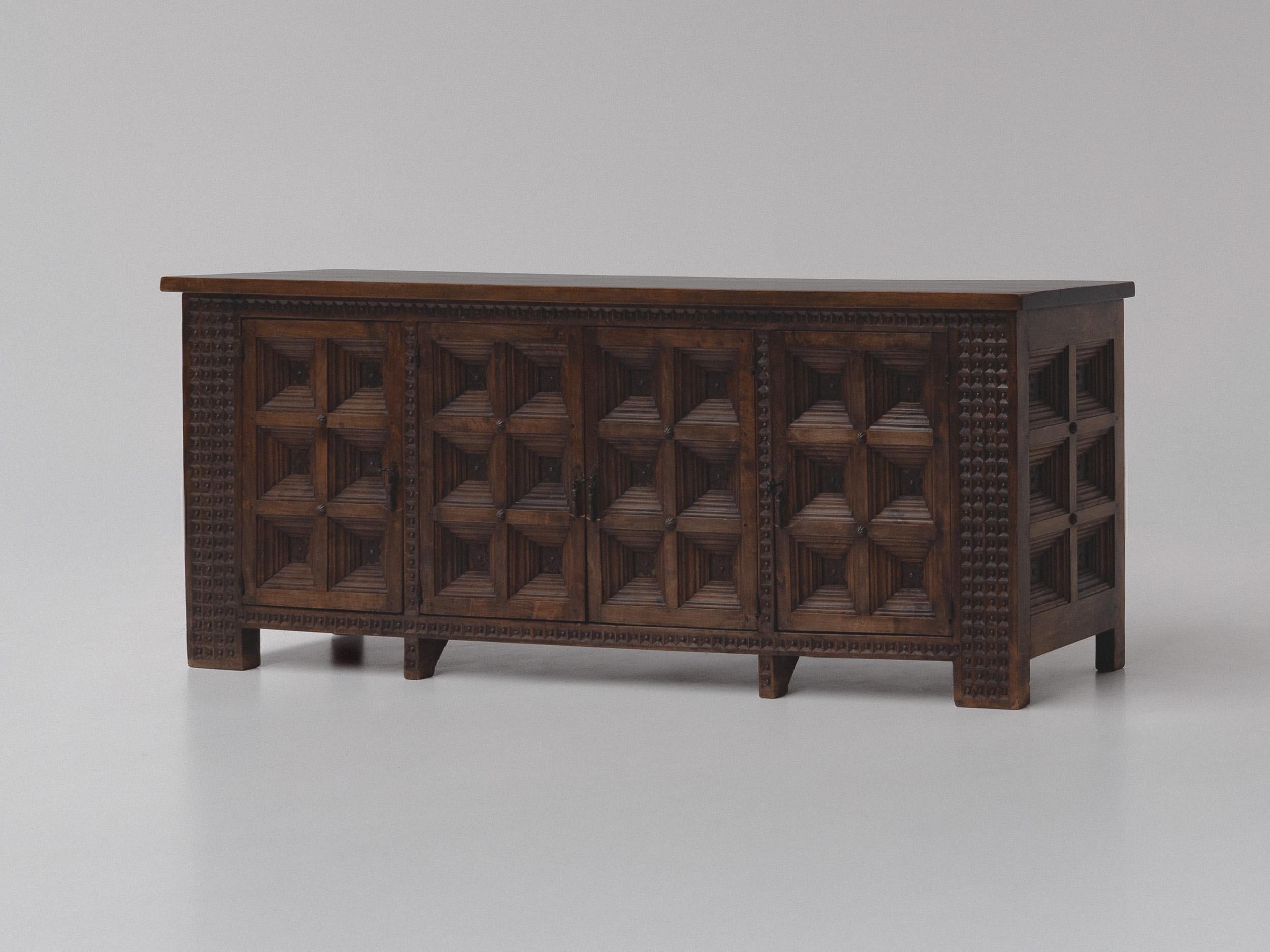 This solid oak Spanish sideboard from the 1960s showcases the timeless charm of rustic craftsmanship. Made from richly grained dark stained pine, the piece features a sturdy construction with bold, geometric paneling on the doors, creating a strong