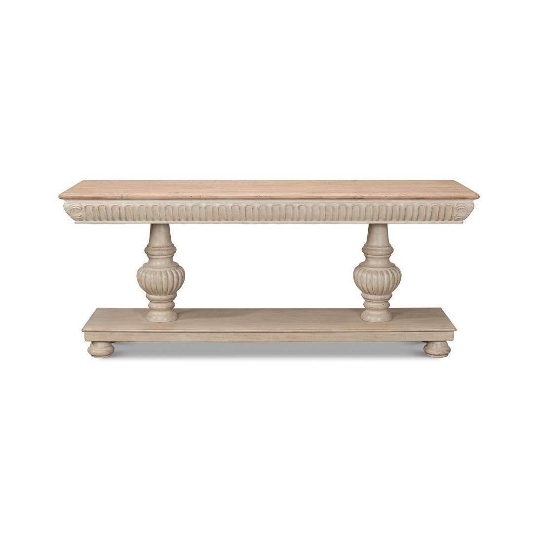 A dramatic transitional pine console, with a natural wooden top above an antiqued stone gray painted base. It features carved volutes around the frieze, atop two carved baluster pedestals. This console table not only serves as a functional surface