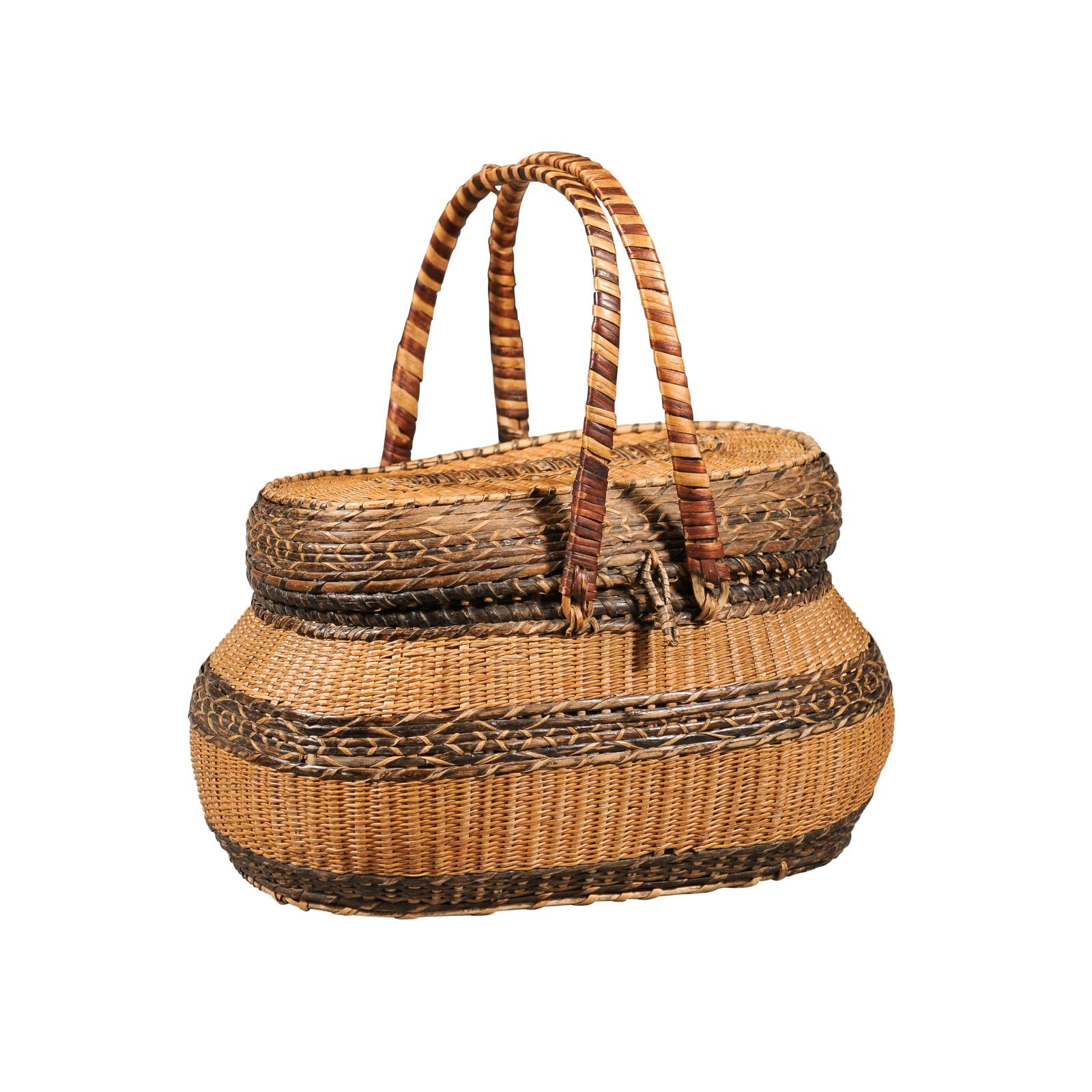 A Swedish oval wicker basket from the late 19th century, with two large handles. Charming us with its rustic appearance and complimenting colors, this Swedish basket features an oval lid opening thanks to a woven latch to reveal a convenient storage