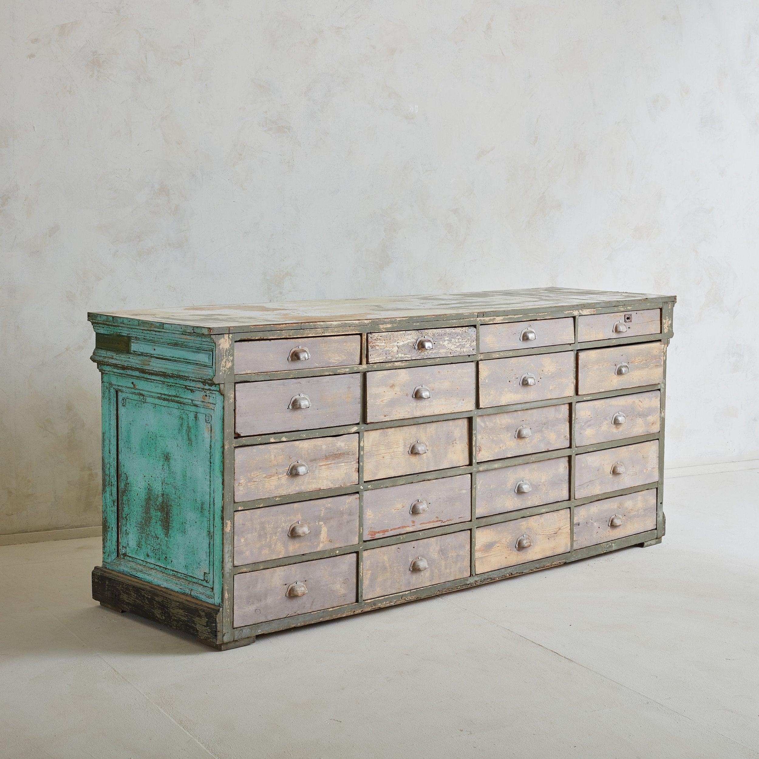 A Rustic painted wood chest of drawers from the 20th Century. This distressed cabinet features 20 drawers, half-round metallic metal pulls, and panel detailing. The painted sides and back have worn to a patina of turquoise and pale blue, contrasting