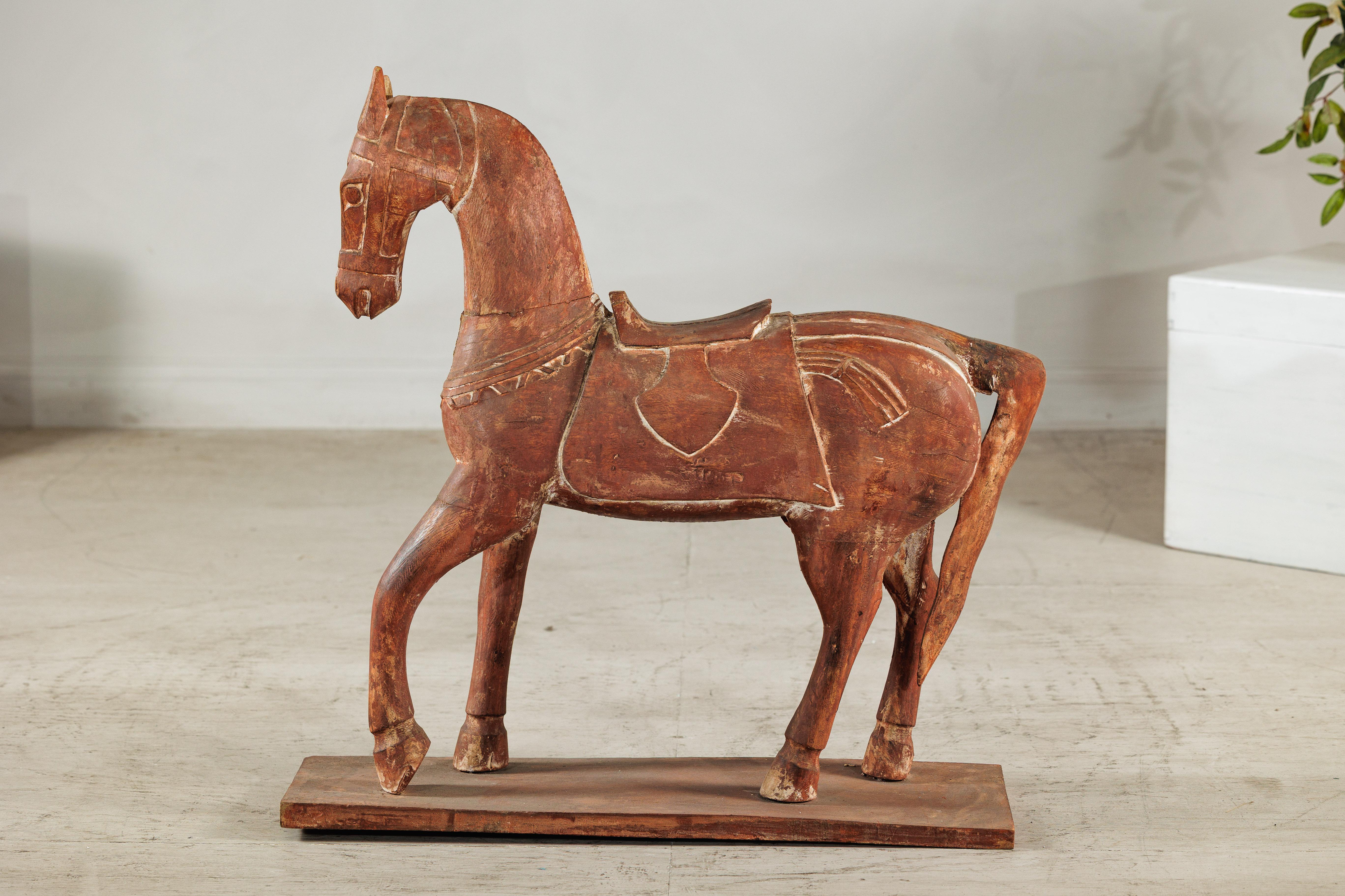 Una scultura di cavallo rustica in legno d'epoca, presentata su una base piatta e ornata da una sella intagliata e da dettagli dei finimenti. La figura è scolpita a mano con una qualità stilizzata, quasi da arte popolare, le sue gambe allungate e la