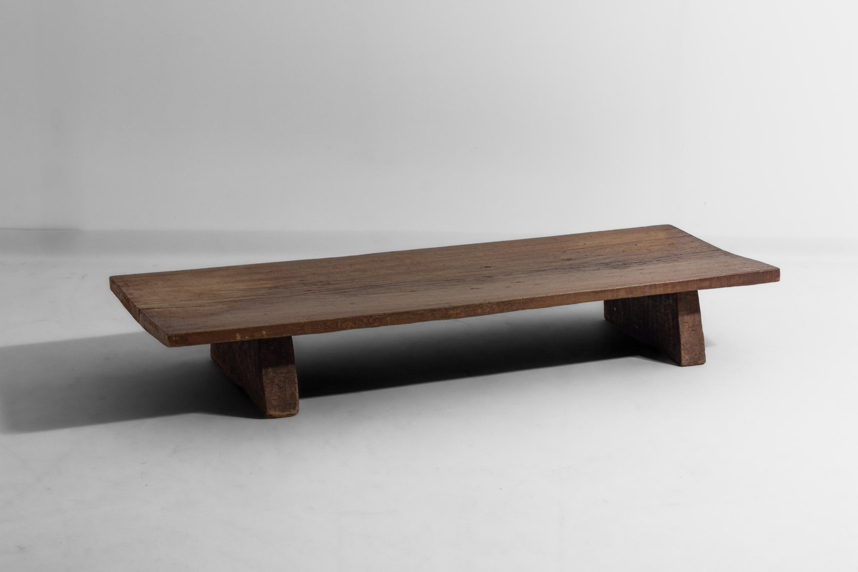 Tavolino in legno massiccio con uno spesso piano intagliato a mano che poggia su due ampie gambe a blocco. La superficie presenta segni di usura e irregolarità tipiche dell'età e dell'uso. Il tavolo ha un aspetto semplice e primitivo con una