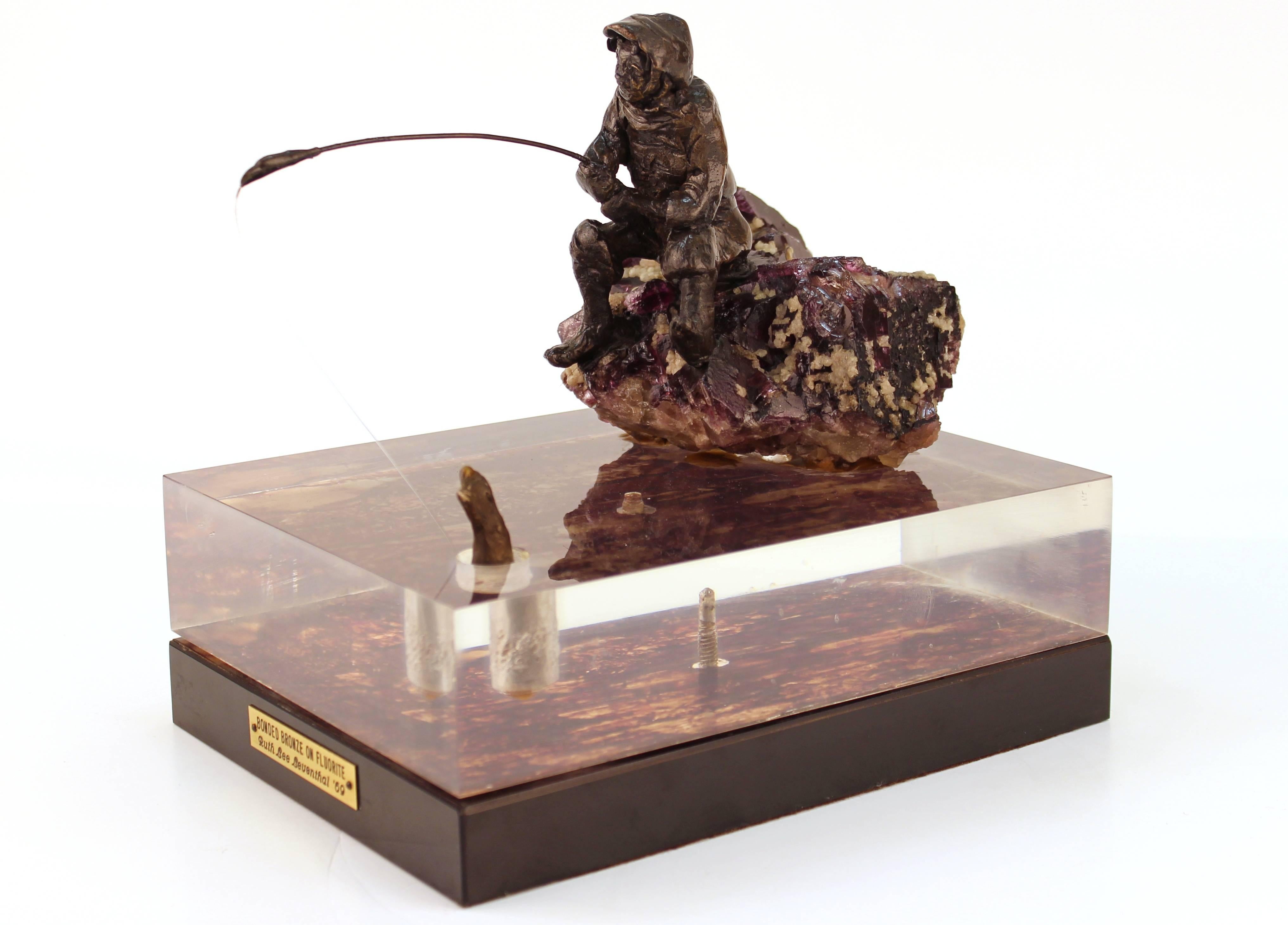 Ruth Lee Leventhal sculpture of a bronze fisherman sitting on a fluorite rock. The fisherman's line drops into a clear Lucite lake with a fish on the other end. The fish piece can sit in our out of the Lucite lake. Includes plaque on base that reads